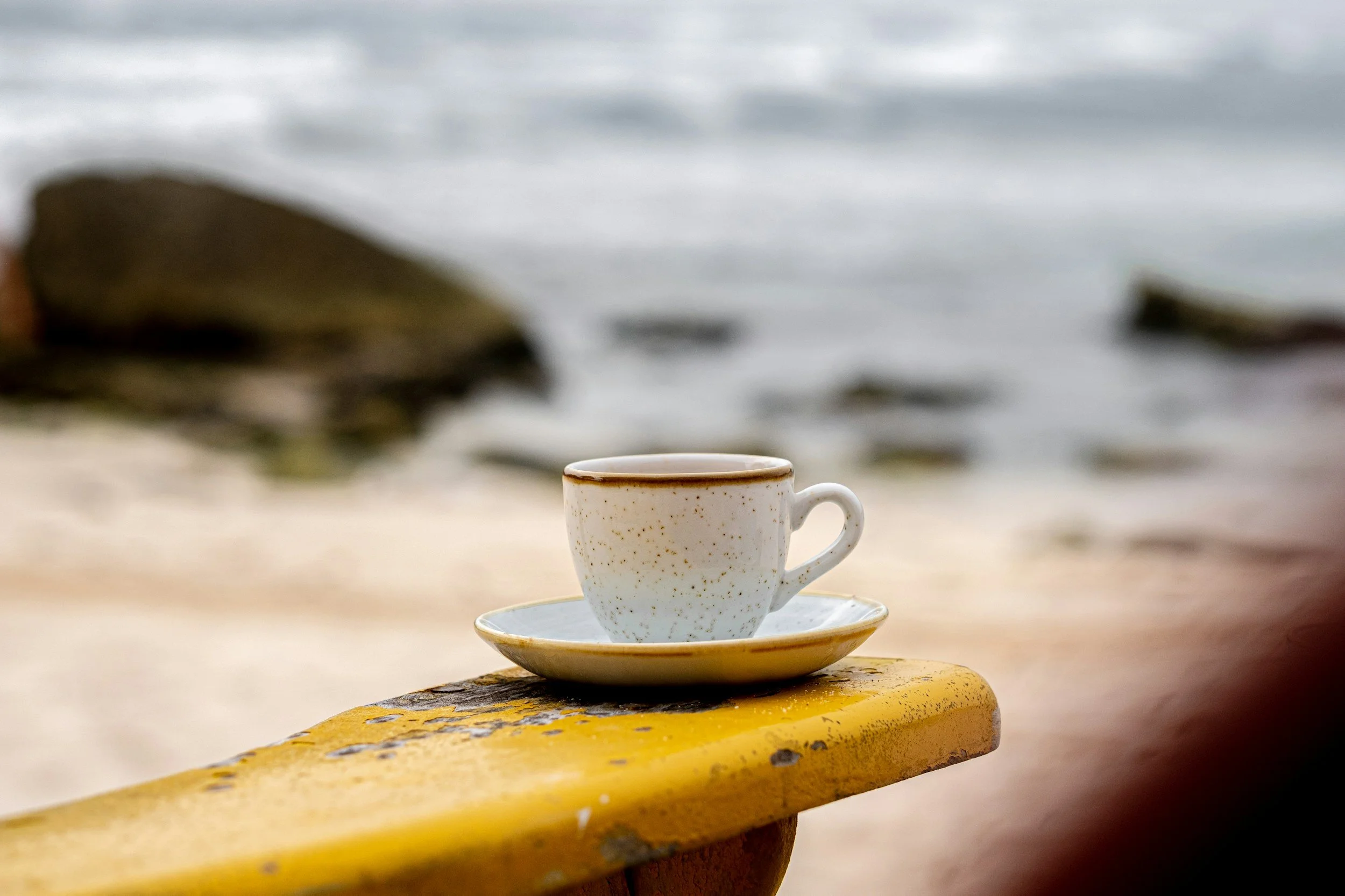 A cup of coffee on a saucer placed on a yellow bench at the beach with rocks and ocean in the background.