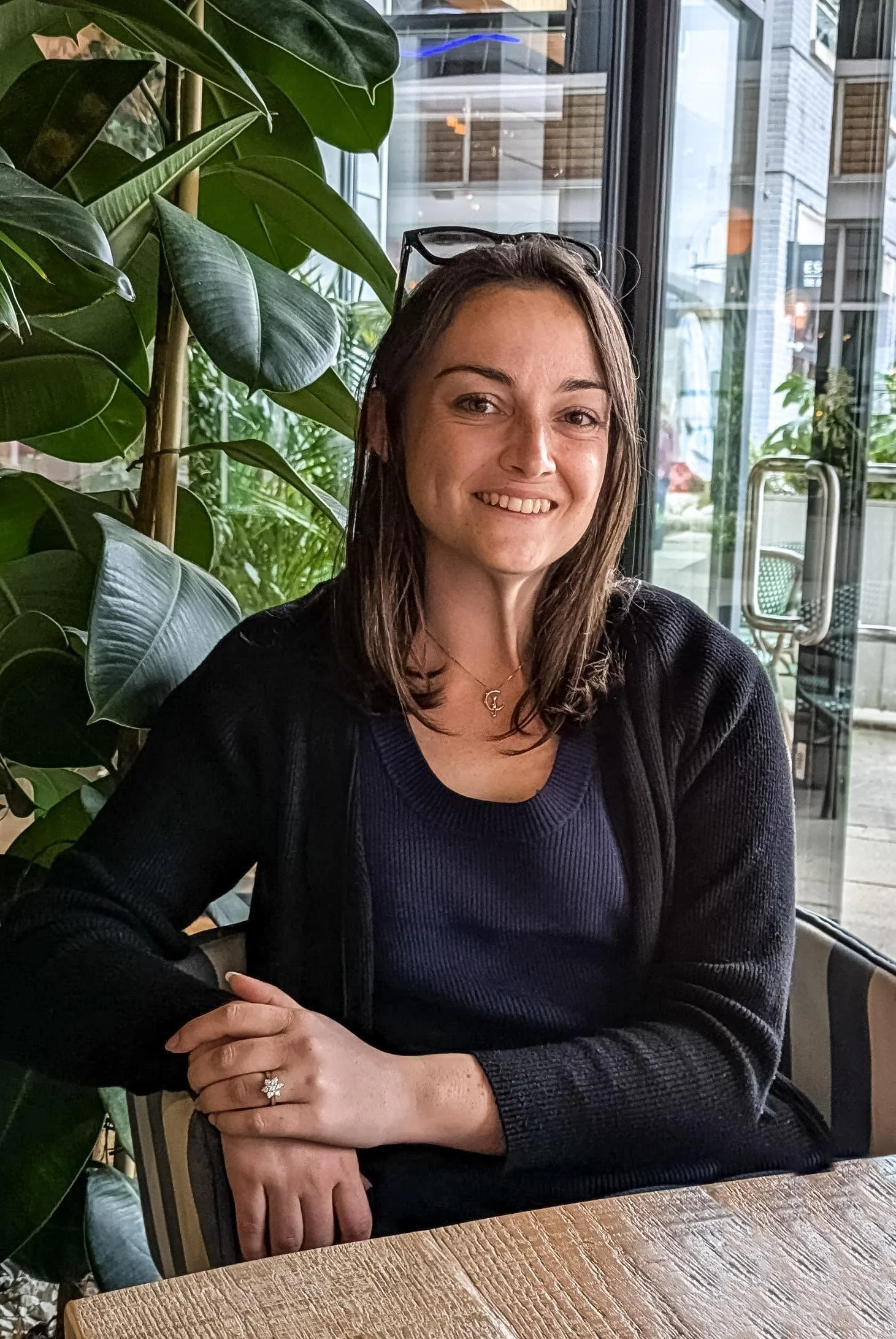 A woman with shoulder-length brown hair, wearing a dark blue top and black cardigan, sitting at a wooden table inside a cafe with large glass windows and green indoor plants.