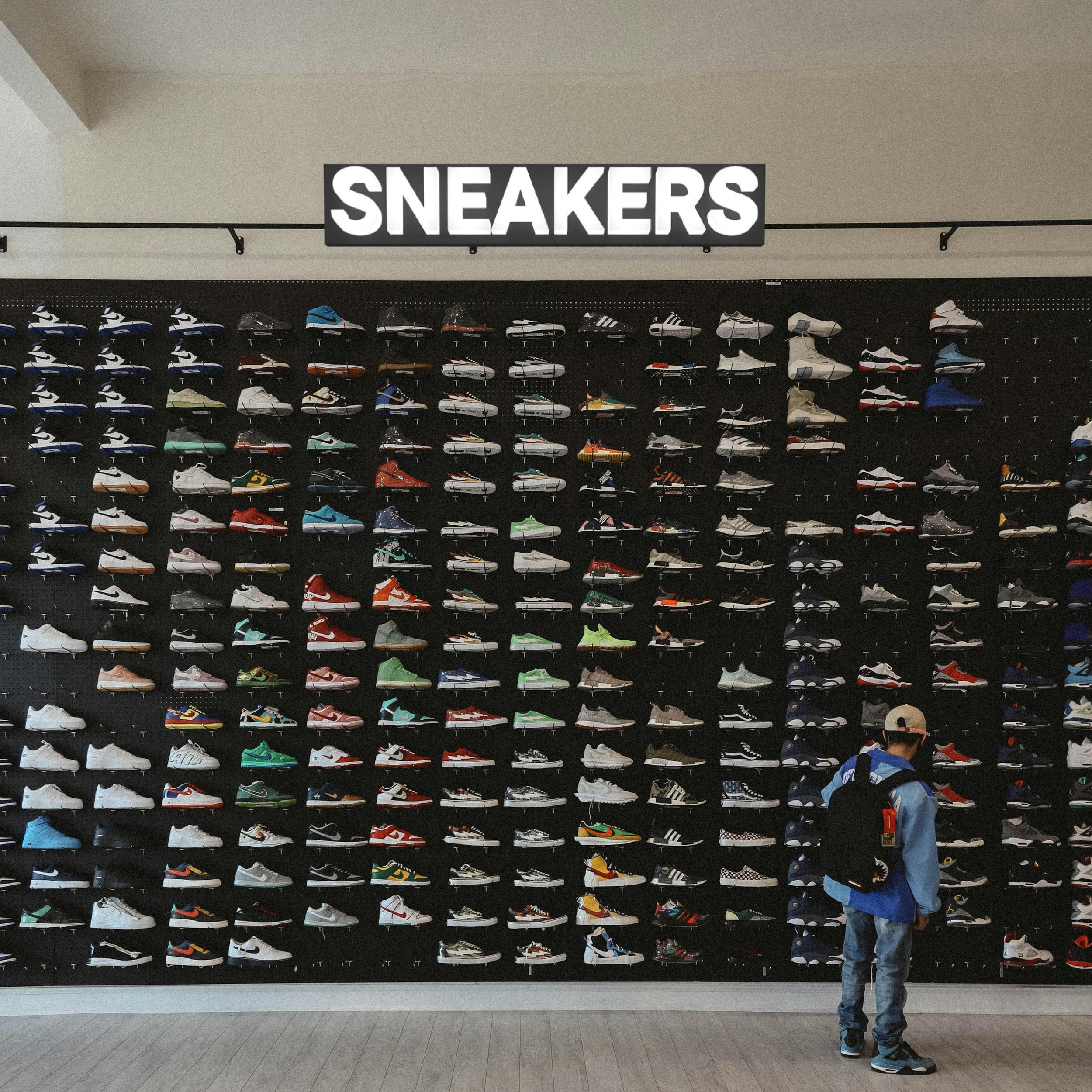A wall display of various sneakers arranged in rows under a sign that says 'SNEAKERS'. A person with a backpack, cap, and jacket looks at the sneakers.