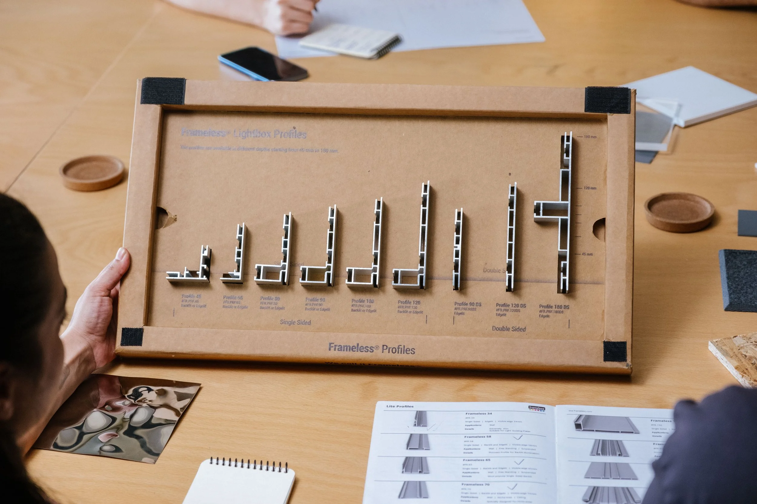 A person holding a cardboard box containing metal profiles arranged in increasing size, labeled with different profiles from 45 to 180, with some labeled 'Single Sided' and others 'Double Sided,' on a wooden table among various papers, a photograph, a smartphone, and small round dishes.