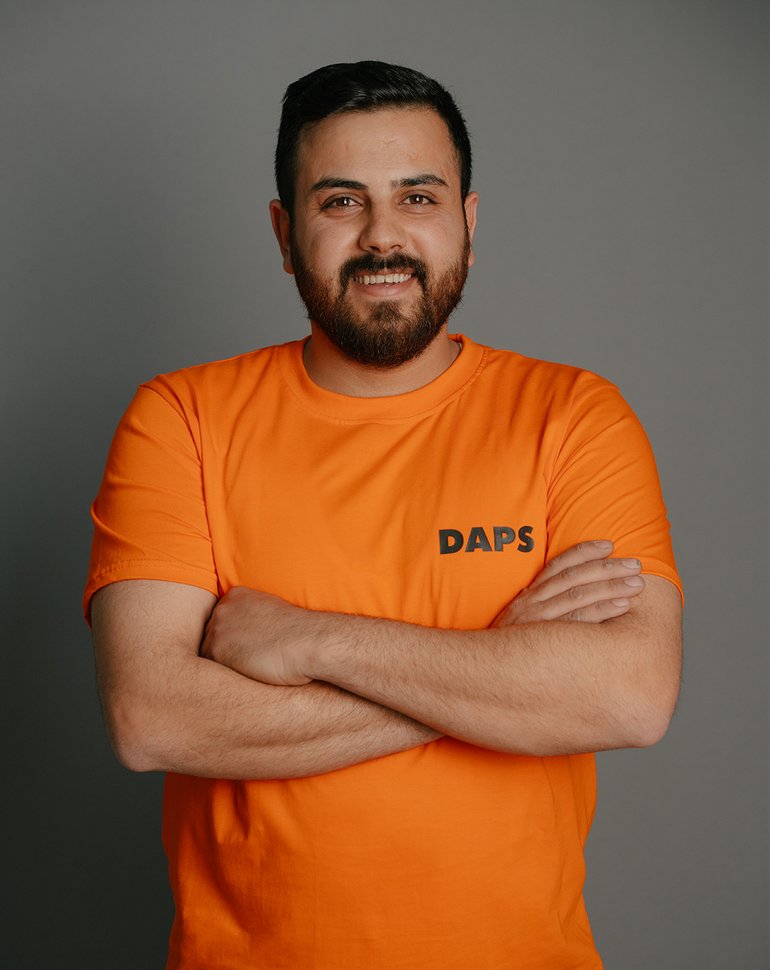 A man with dark hair and a beard smiling with crossed arms, wearing an orange t-shirt with 'DAPS' written on it, standing against a gray background.