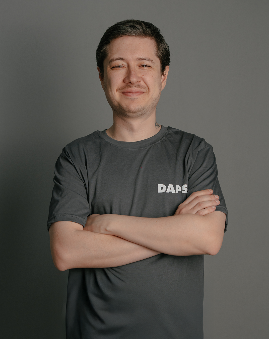 A young man with short dark hair, light skin, and a slight smile standing with arms crossed against a gray background, wearing a dark gray T-shirt with the word 'DAPS' printed on it.