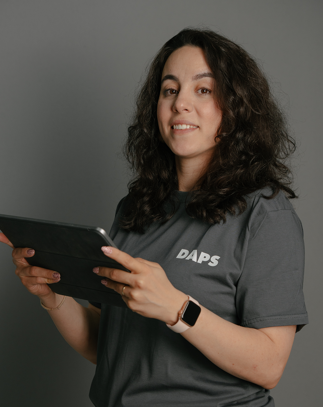 A woman with long, curly dark hair wearing a dark gray T-shirt with the word 'DAPS' on it, holding a tablet and smiling at the camera against a gray background.