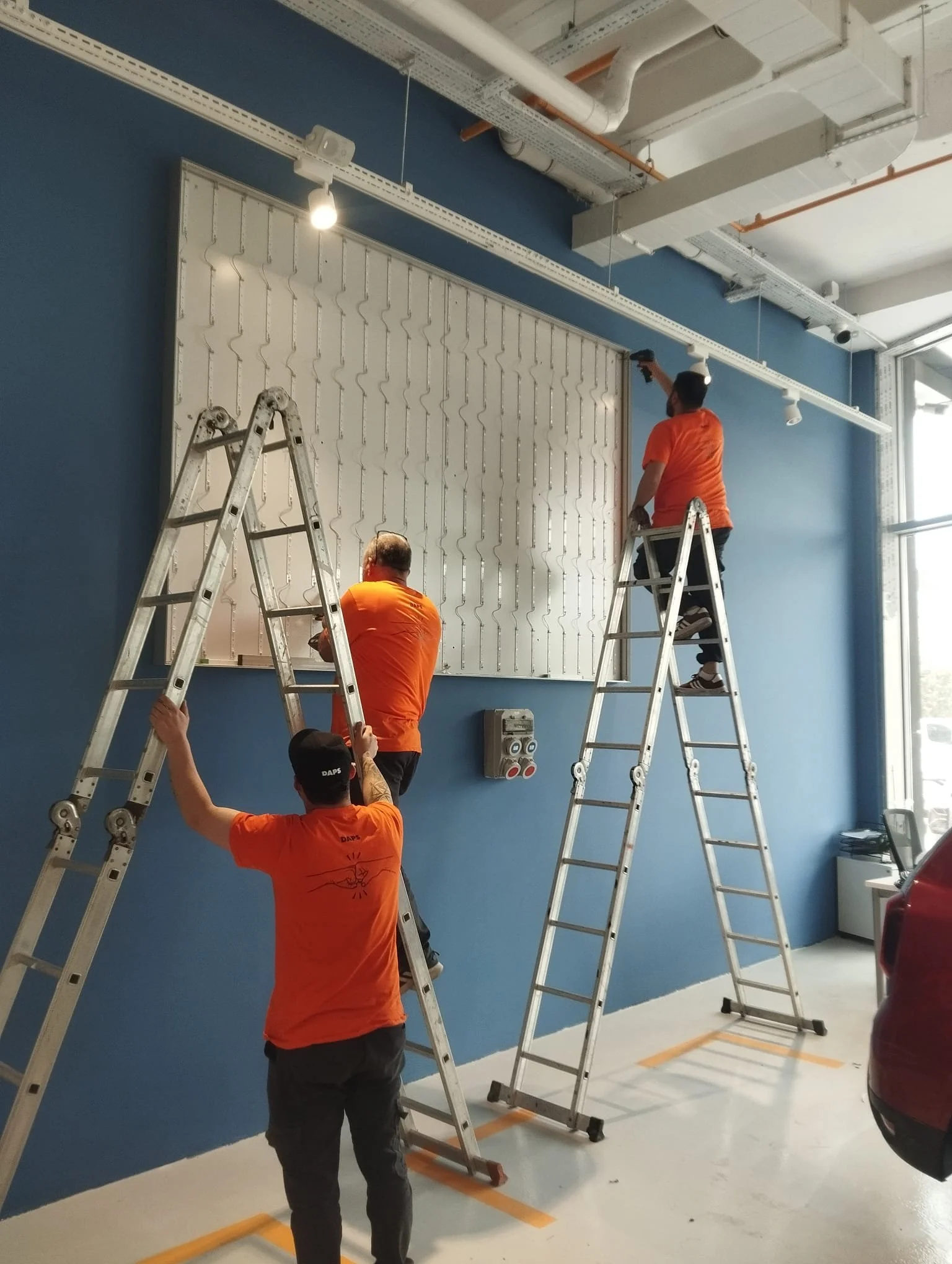 Three workers in orange shirts and black pants installing or repairing equipment on a whiteboard or display panel on a blue wall in an indoor space, using ladders.