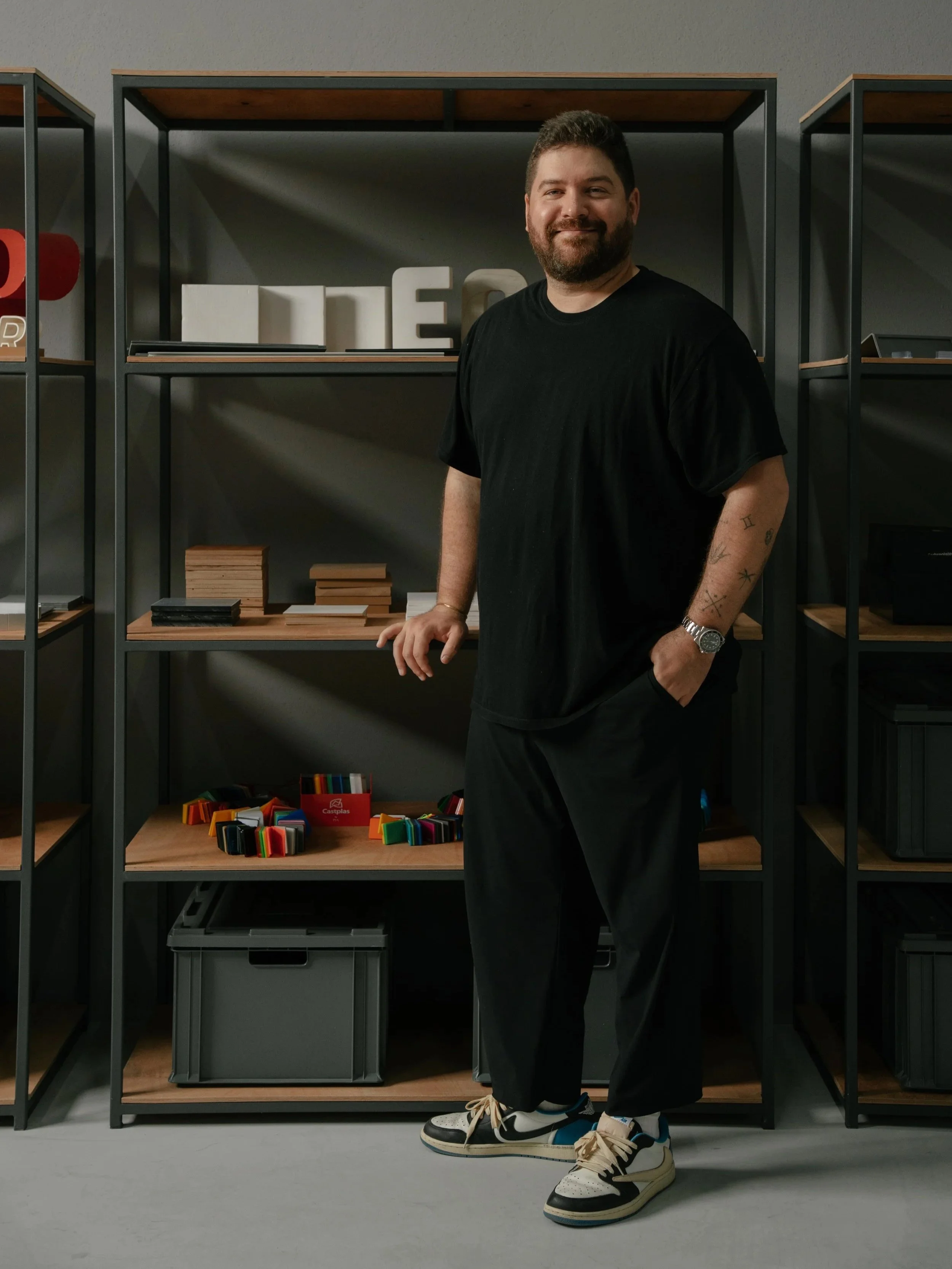 A man with a beard and tattoos on his arm stands in front of shelves with books and colorful objects, smiling at the camera.