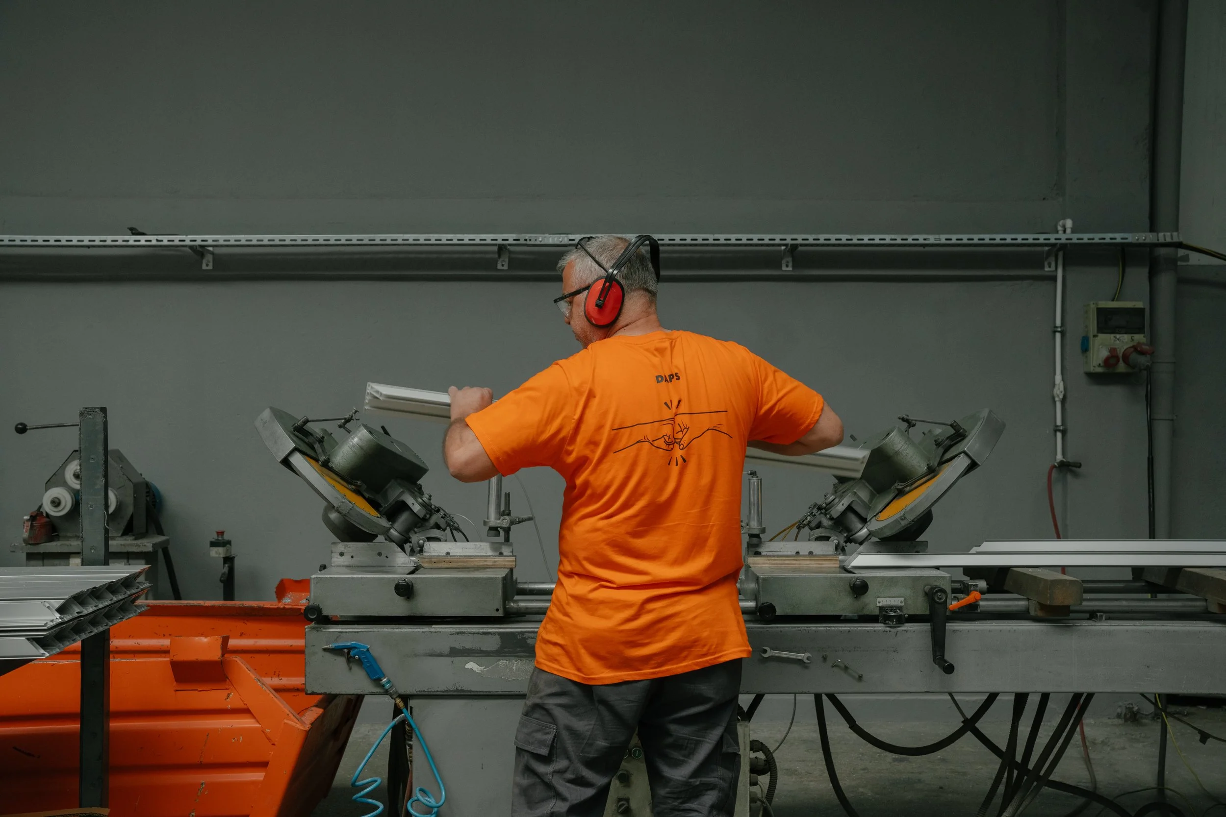 A worker with gray hair, glasses, and a beard wearing an orange t-shirt and gray pants is working at a machine in an industrial setting. The worker is wearing red ear protection and appears to be adjusting or inspecting the machinery.