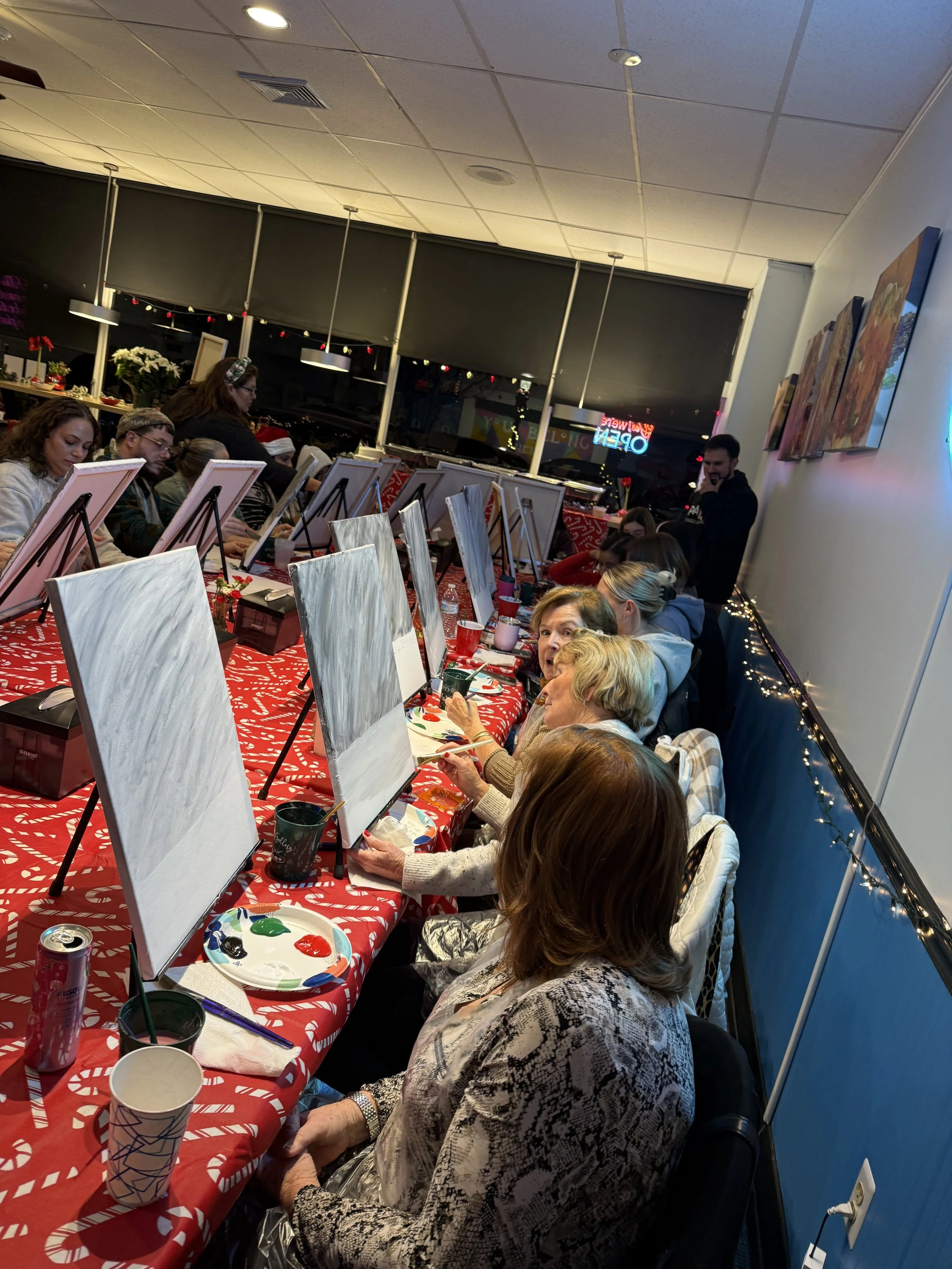 People participating in a group painting activity at a long table with Christmas decorations, using canvases and paints, in a well-lit indoor space.