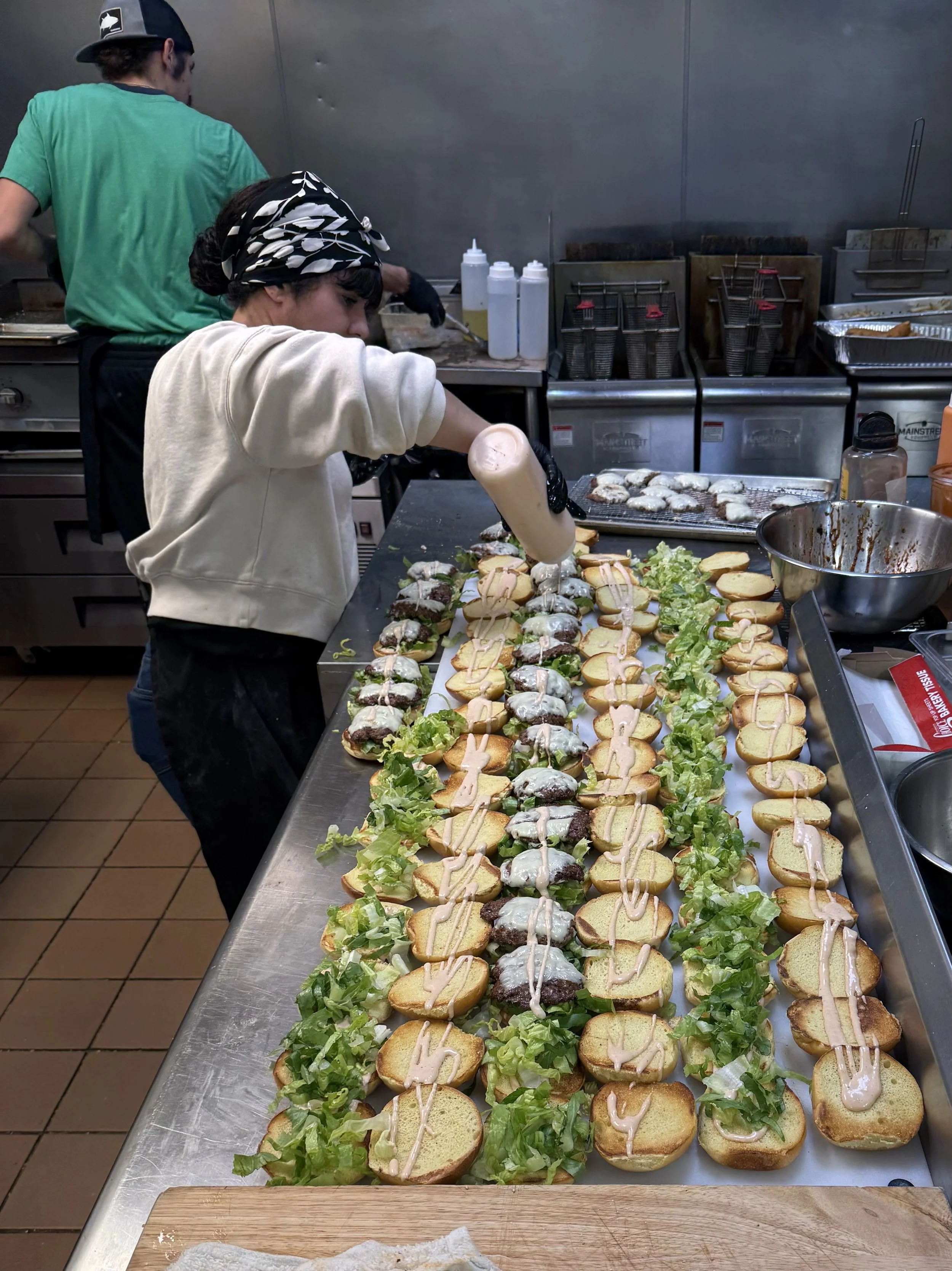 A worker prepares multiple cheeseburgers with lettuce on a stainless steel counter in a commercial kitchen.