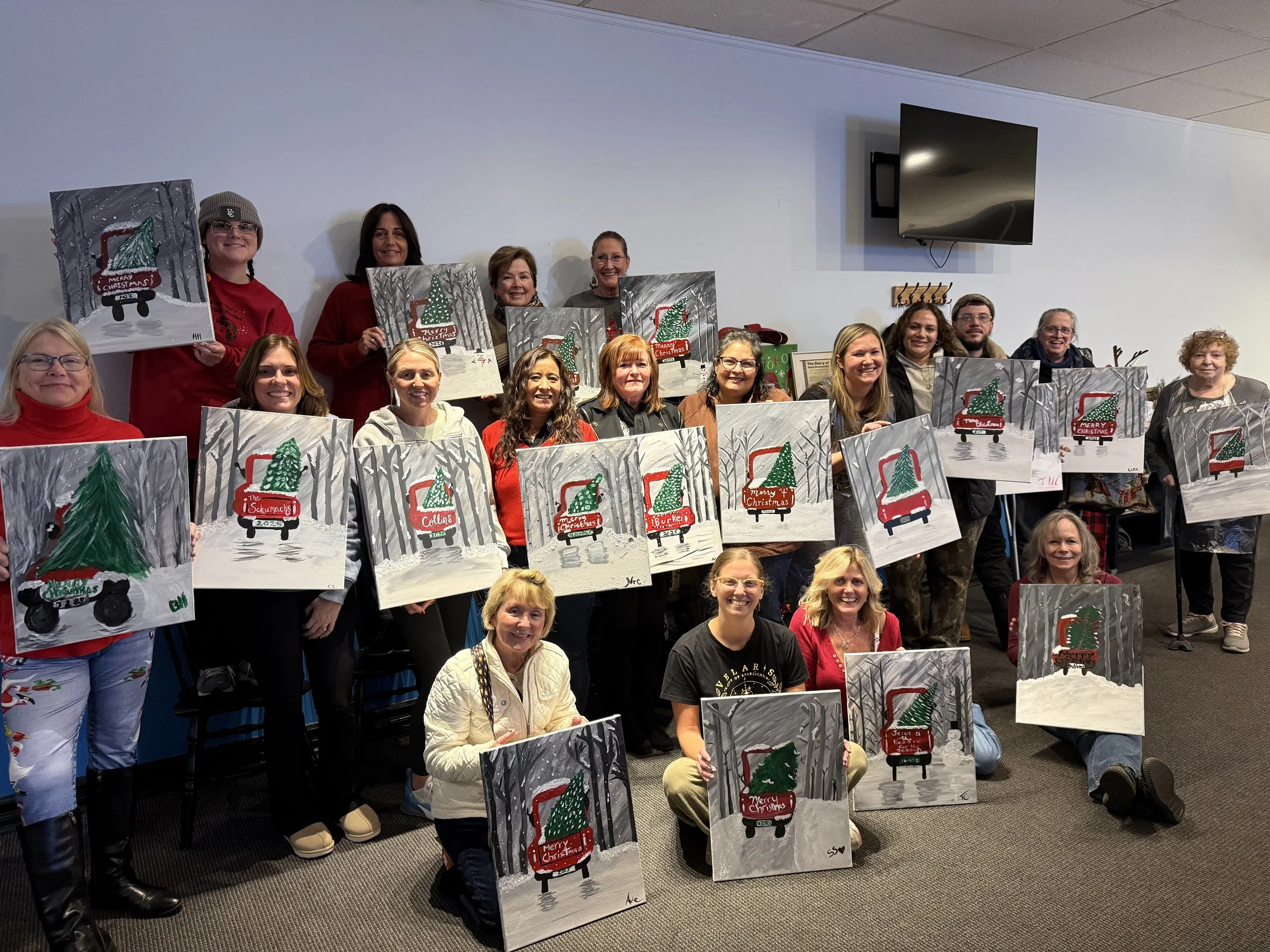 Group of people holding Christmas-themed paintings of a Christmas tree on a wagon with the text "Merry Christmas."