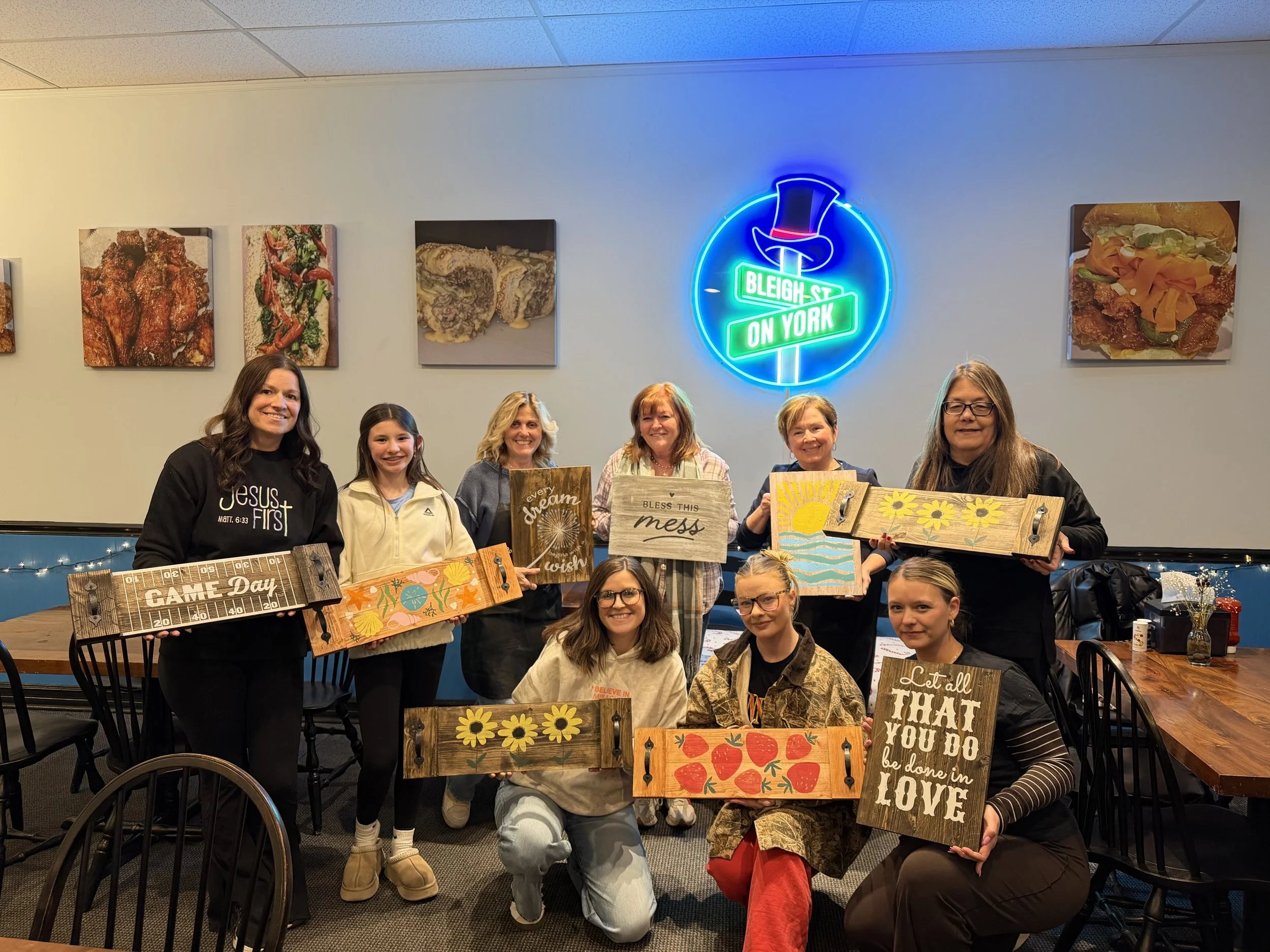 Group of ten women and girls standing together inside a restaurant, holding decorated signs with inspirational and seasonal messages. The restaurant has a neon sign that reads "Bleak Street on York" and food photographs on the wall.