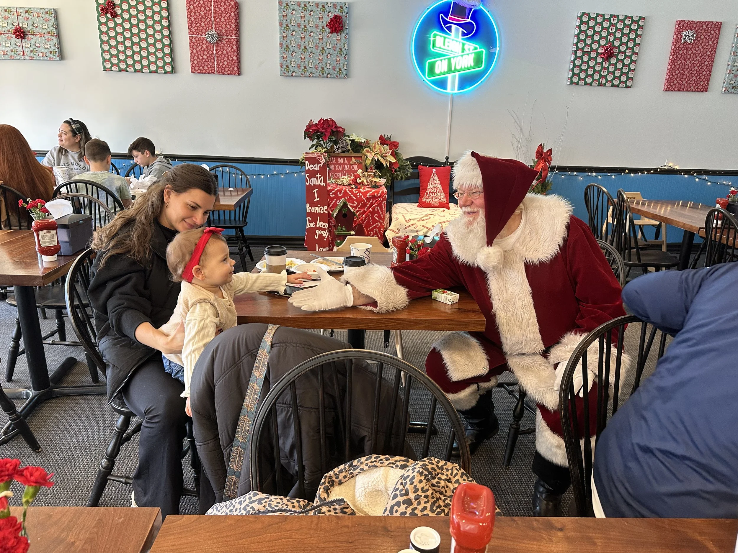 Santa Claus dressed in a red suit with white fur trim sitting at a table in a restaurant, greeting a young girl with a woman sitting beside her. The girl has a red headband and beige sweater. The restaurant is decorated with Christmas ornaments and presents, and there are other families in the background.