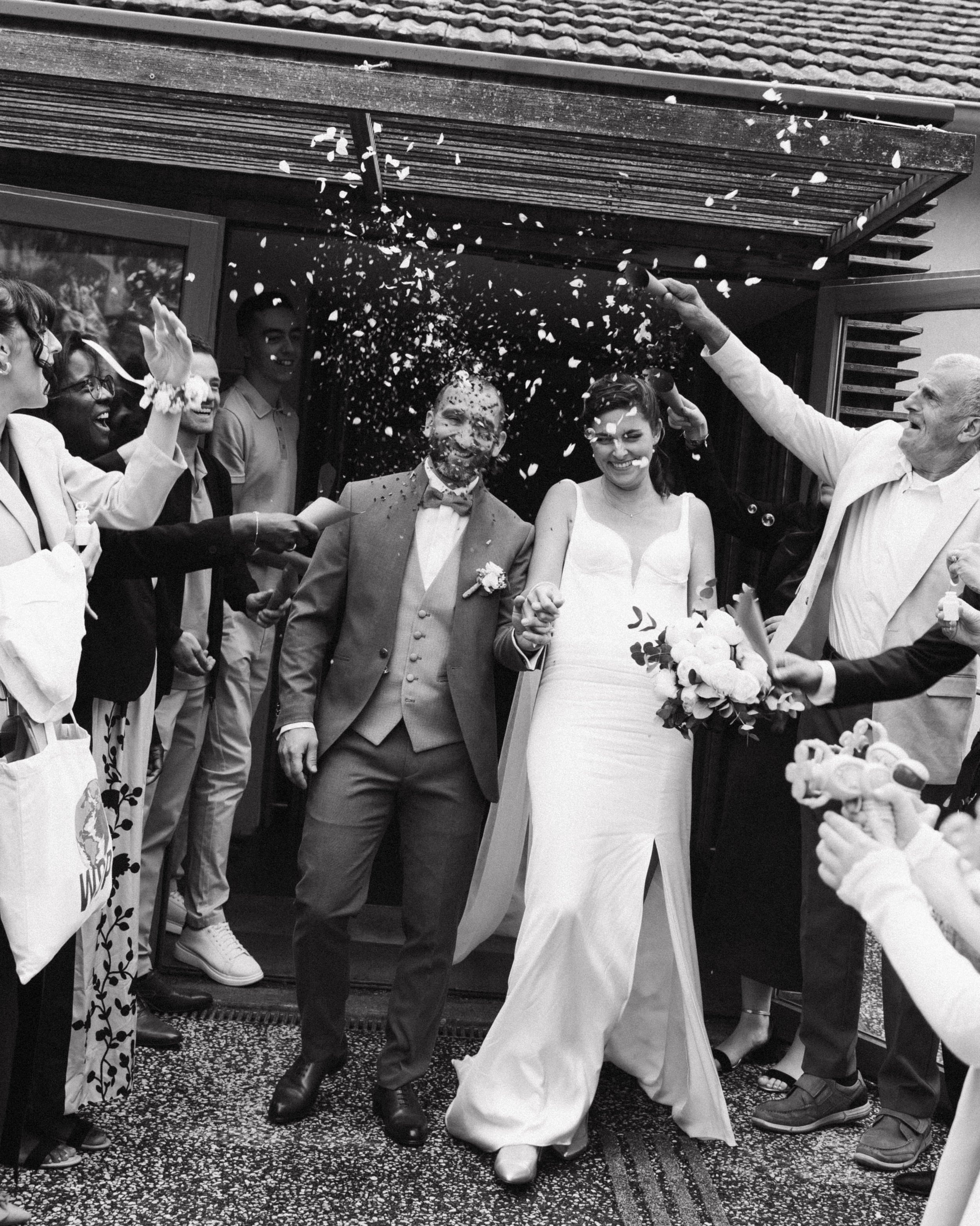 Un groupe de personnes en costume et habillées élégamment, célébrant un mariage, avec des confettis dans l'air, devant une maison.