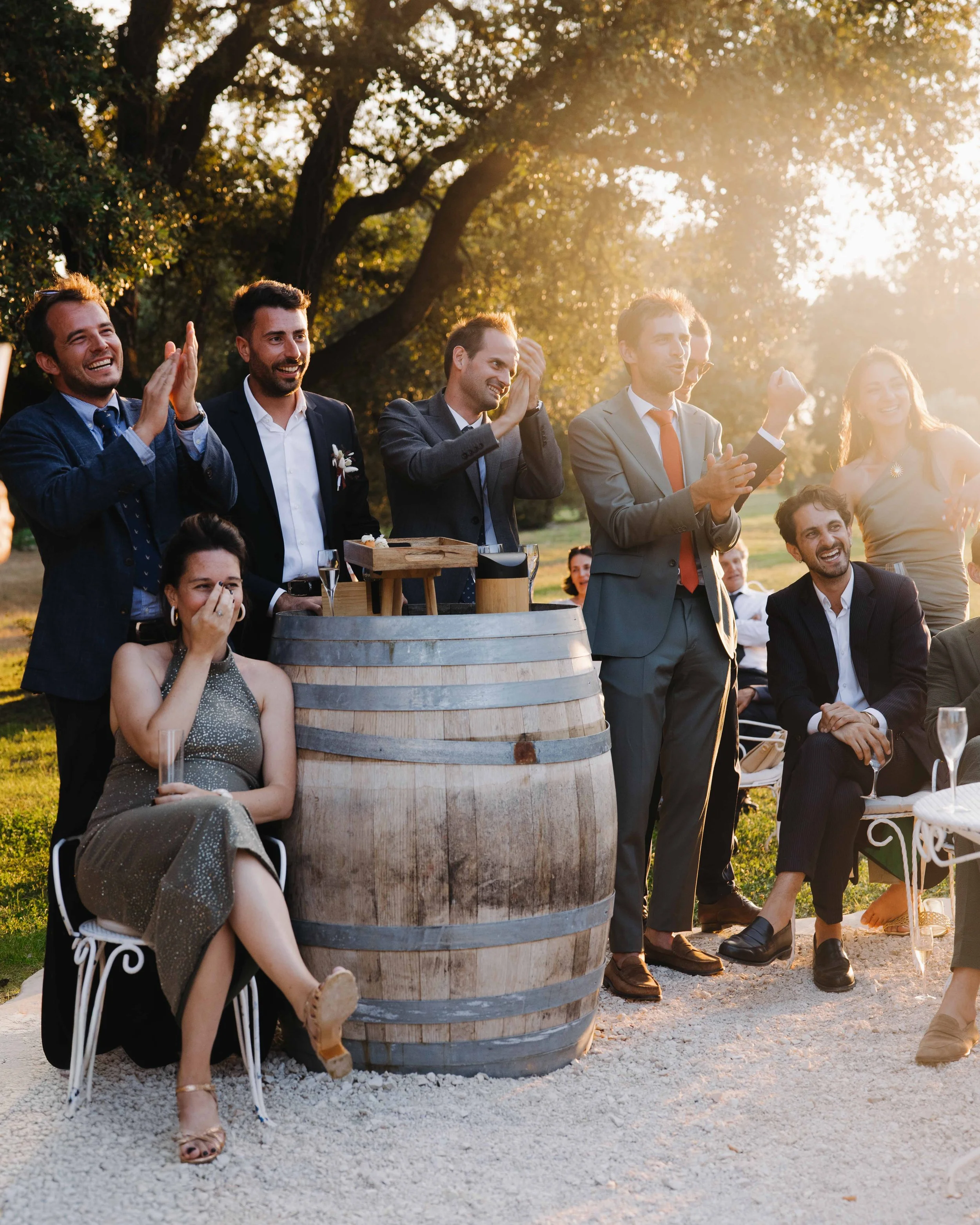 Groupe de personnes parlant et souriant lors d'une fête en plein air, avec un tonneau en bois et des arbres en arrière-plan, en fin d'après-midi.