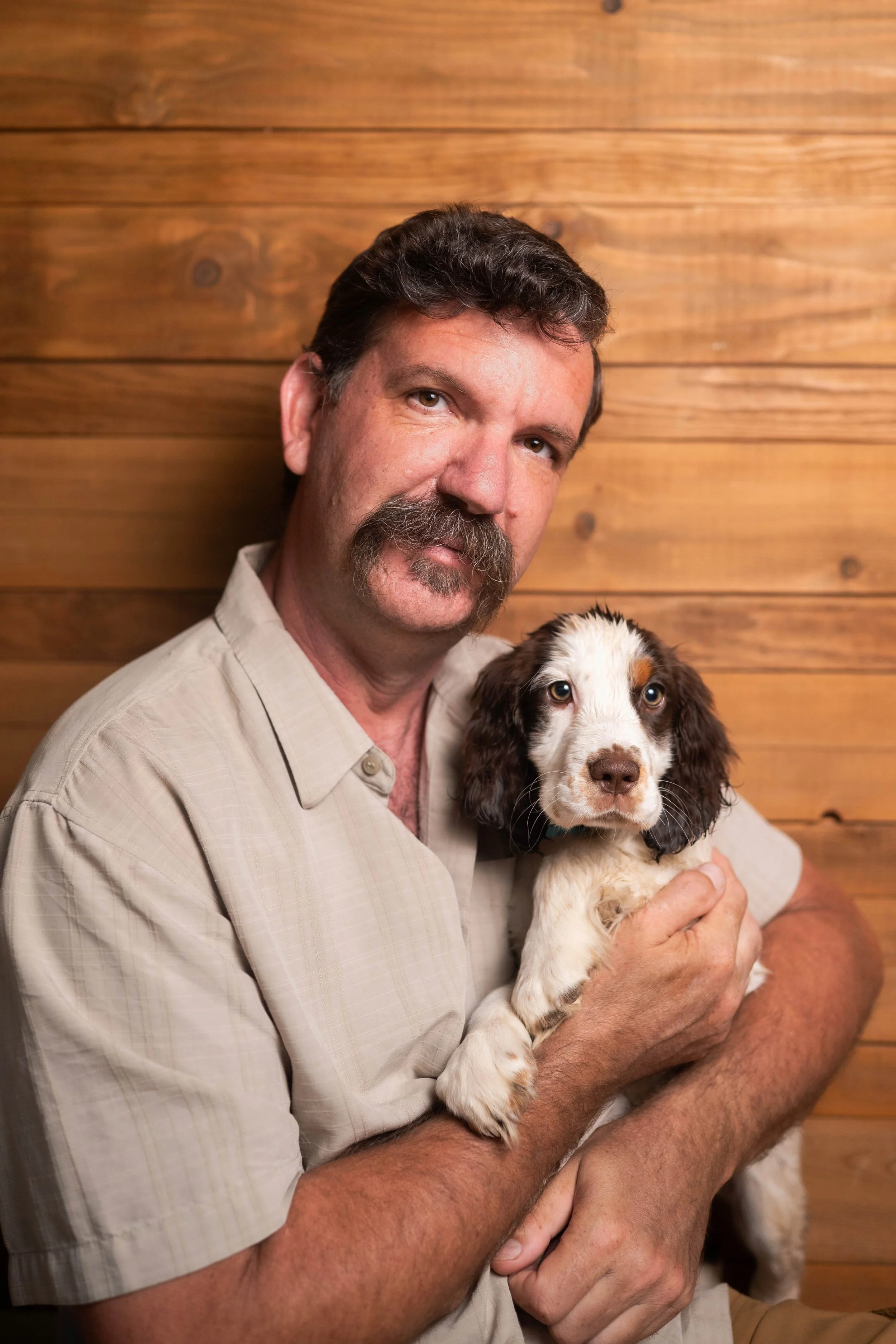 A man with a mustache and beard holding a brown and white puppy in his arms, standing against a wooden wall.