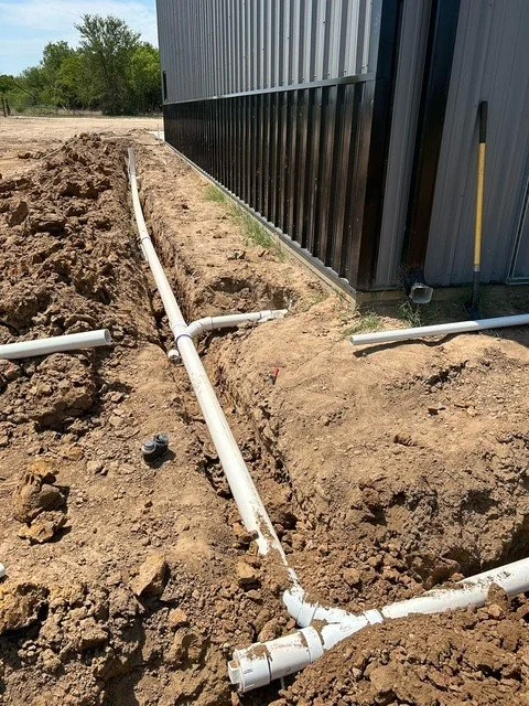 White PVC pipes laid in trenches alongside a building with a black metal exterior, next to a ladder, on a dirt ground with trees in the background.