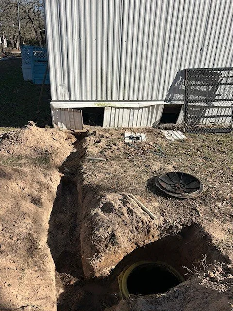 Construction site with a large dirt trench near a metal building, a black pipe, and miscellaneous construction materials.