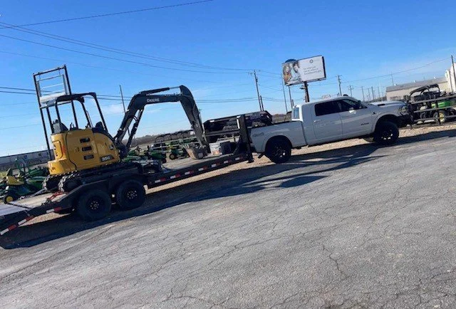 A white pickup truck parked on a gravel lot with a small yellow excavator on a trailer attached to it.