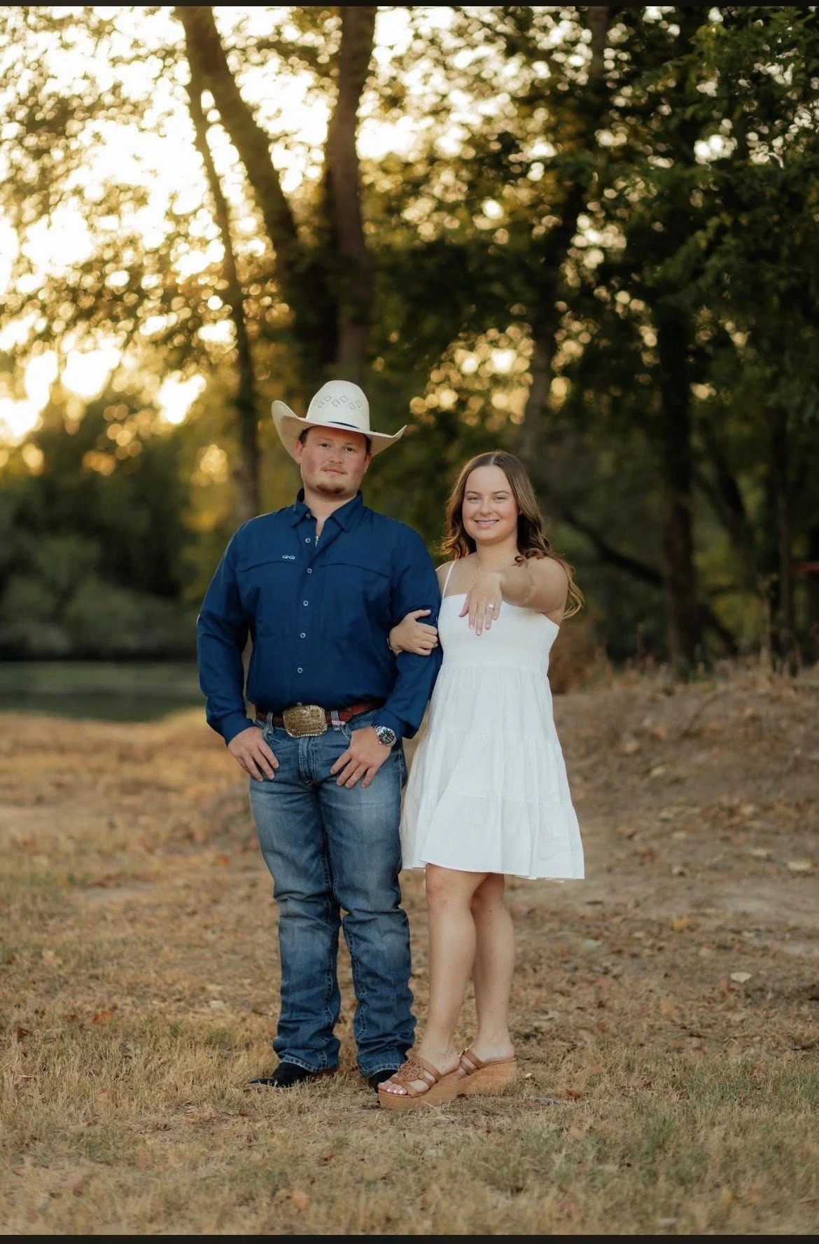 A young man wearing a cowboy hat and blue shirt standing arm in arm with a young woman in a white dress outdoors during sunset.