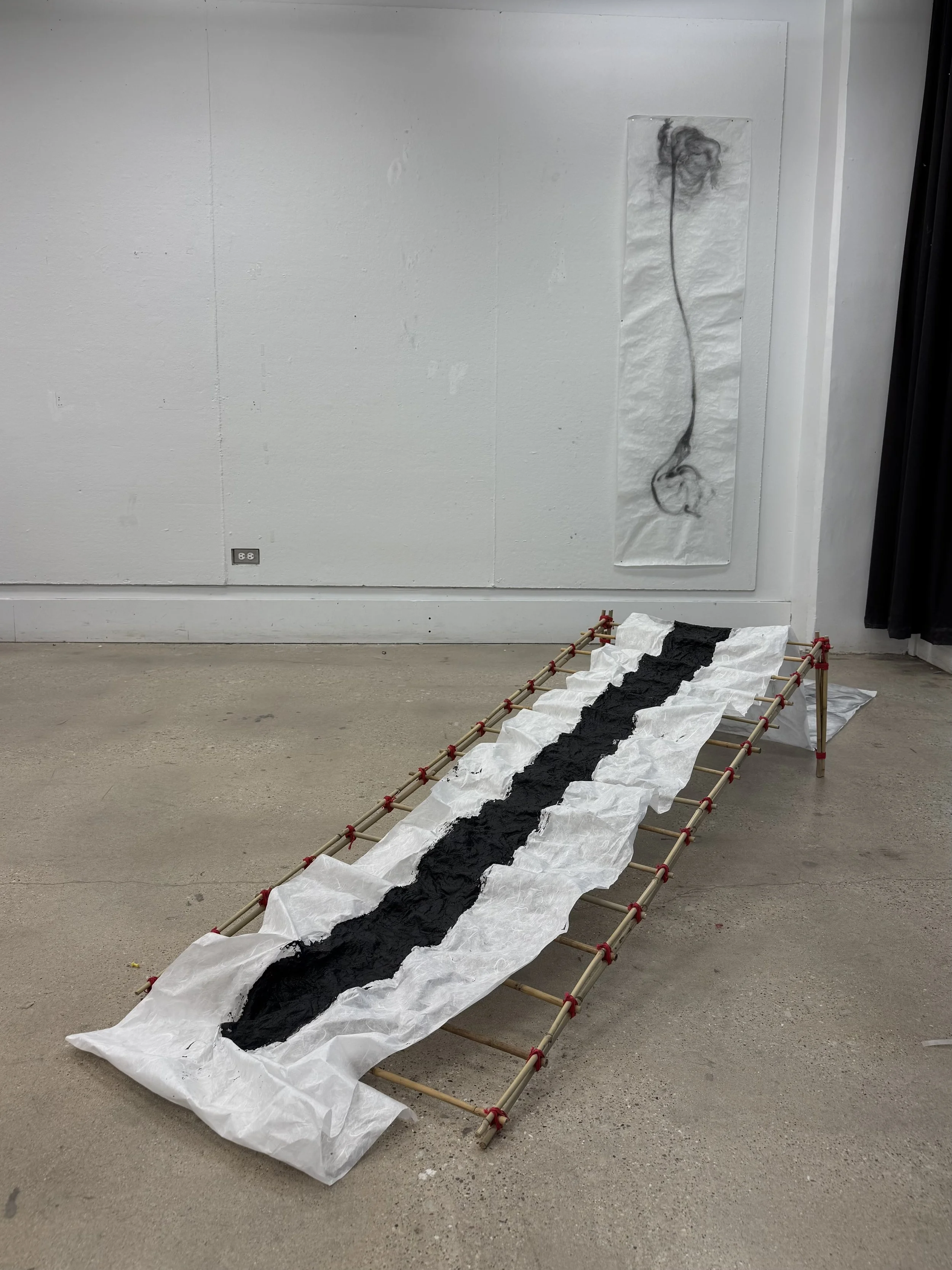 An art installation featuring a fabric sheet with black ink on white paper, laid over a wooden frame structure on a concrete floor in an indoor gallery space, with a large black-and-white drawing of a flower on the wall behind.