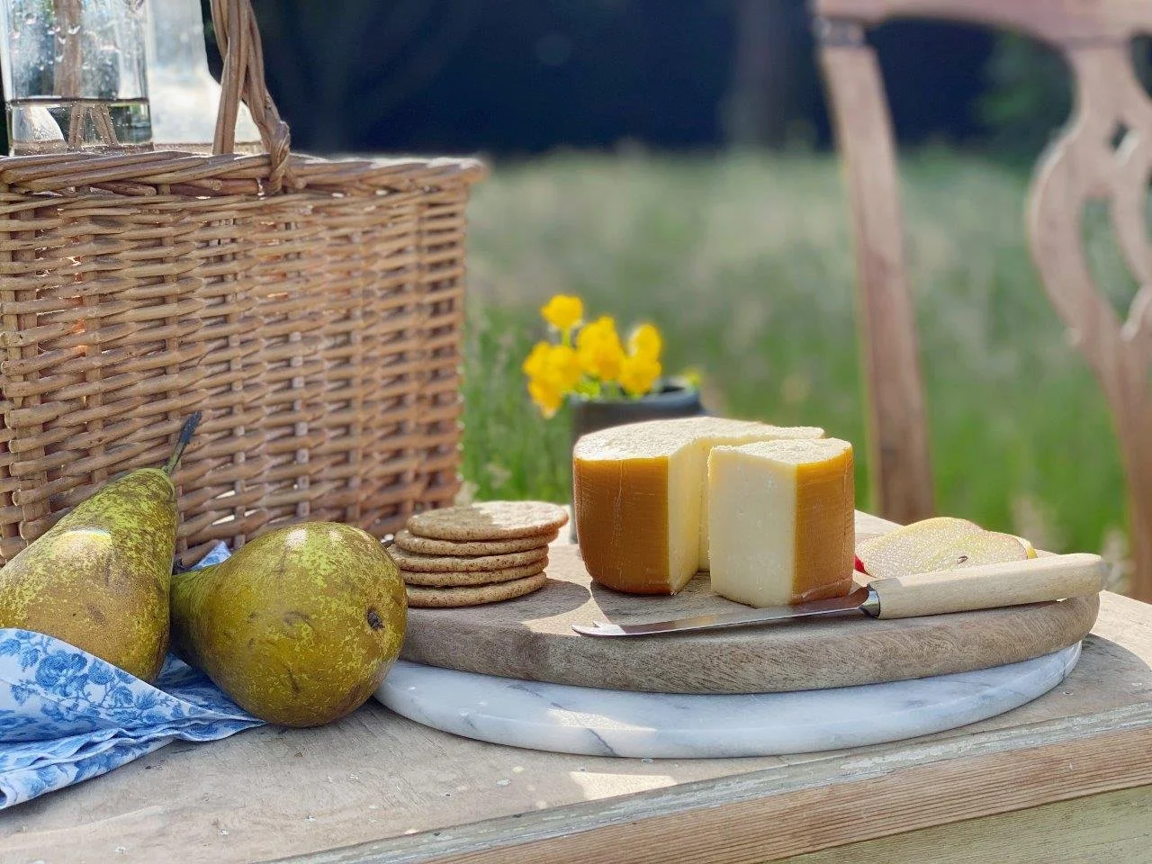 A rustic outdoor setting with two pears, a stack of crackers, and two blocks of cheese on a wooden cutting board. There is a wicker basket and a small pot of yellow flowers in the background.