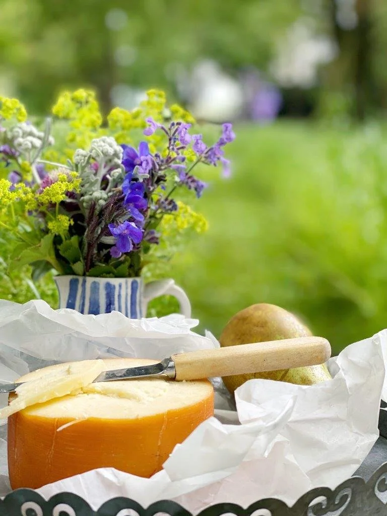 A cheese wheel with a piece cut out, a cheese knife with a wooden handle, a pear, and a purple and yellow flower arrangement in a striped ceramic vase on a table outdoors with a blurred green background.