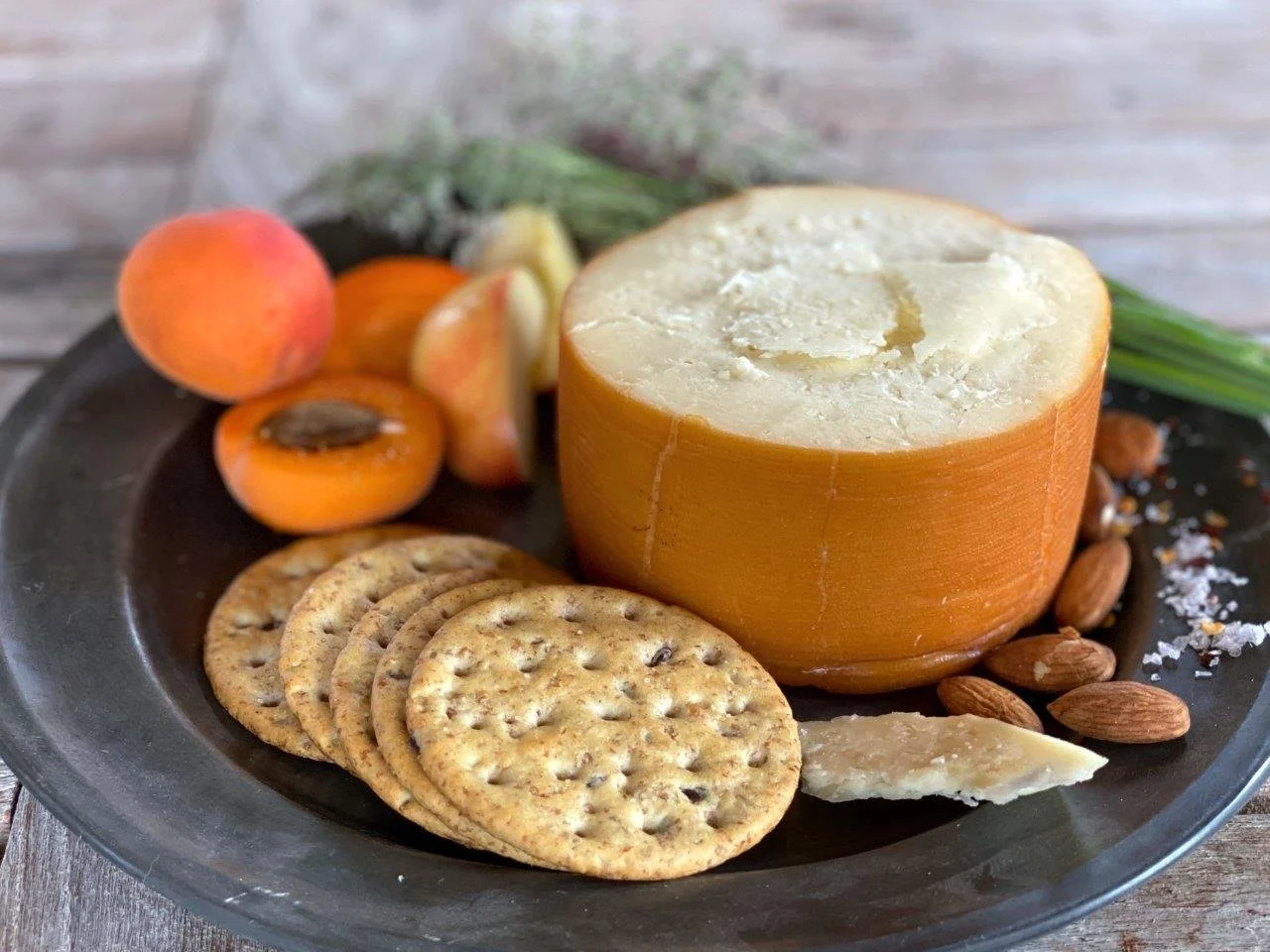 A cheese wheel, whole and cut, with crackers, apricots, almonds, and green onions on a dark plate.