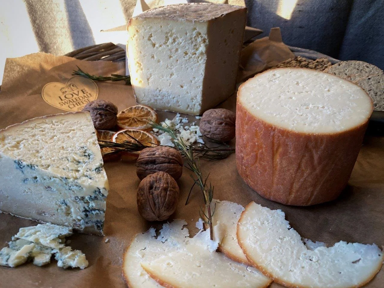 Variety of cheeses including a large wheel of soft cheese, a wedge of blue cheese, and a block of semi-hard cheese, garnished with walnuts, dried lime slices, and sprigs of herbs on parchment paper.
