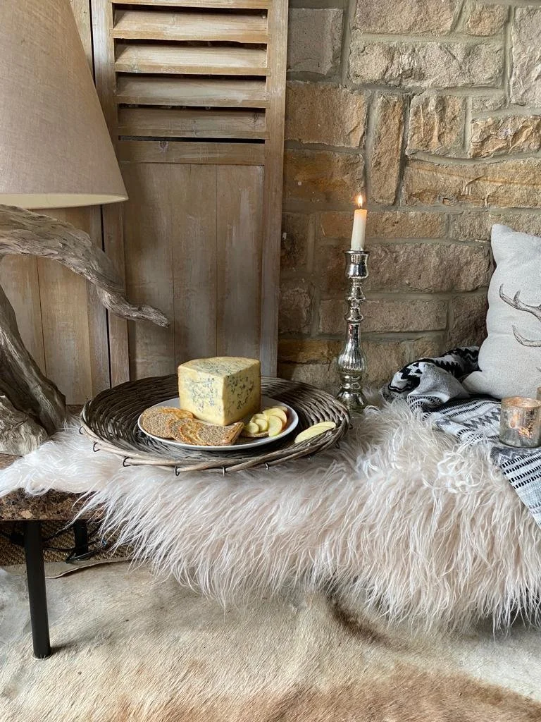 A cozy living room corner with a sheepskin rug, a decorative throw pillow with a deer silhouette, a table with cheese, crackers, and apple slices, a tall candlestick with a lit candle, and a wooden wall and brick background.