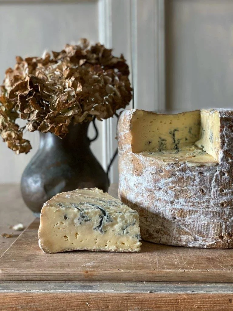 A piece of blue cheese and a block of blue cheese with mold on a wooden cutting board, with a dried hydrangea flower in a vase in the background.