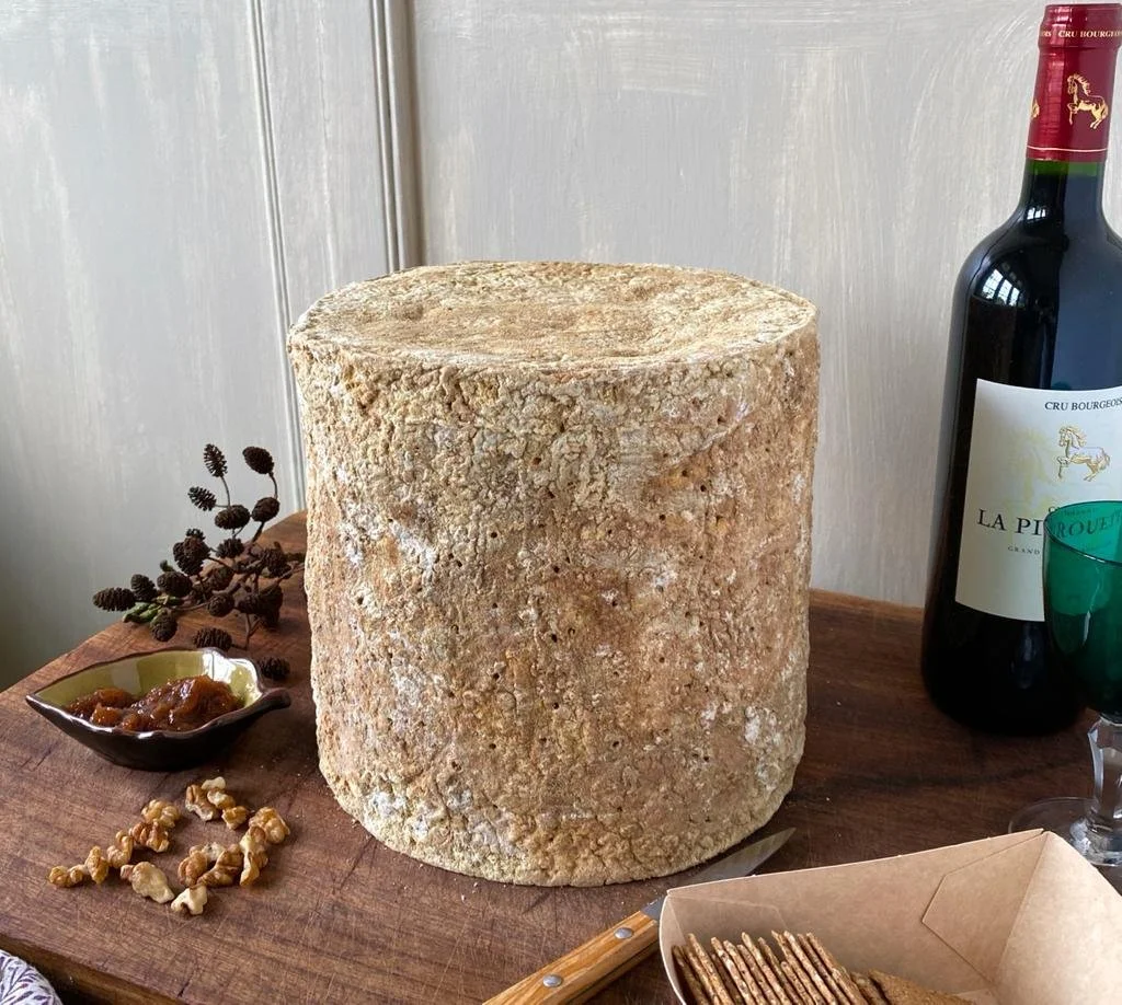 A large wheel of aged cheese on a wooden serving board with a knife, alongside a small dish of jam, walnuts, and dried berries, bottles of red wine, and a green wine glass.