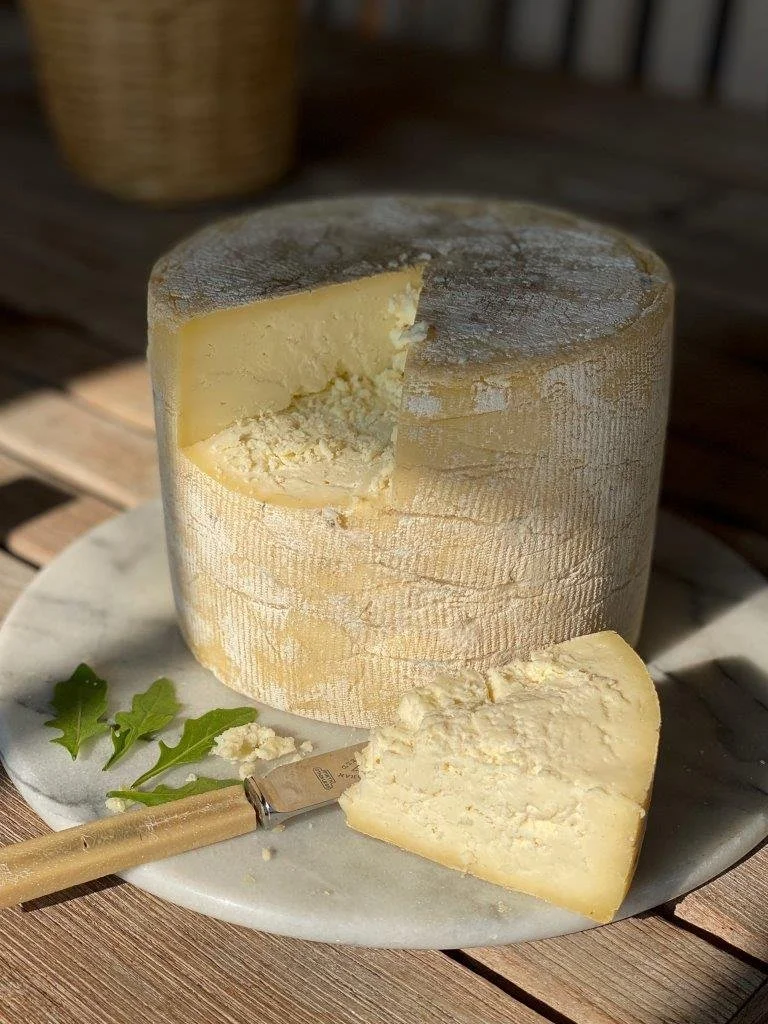 A round wheel of hard cheese with a wedge cut out, placed on a marble slab with a cheese knife and some green leaves for garnish.