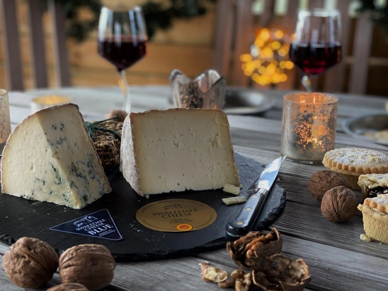 A cheese platter featuring blue cheese and Swiss cheese on a black slate board, surrounded by walnuts, pecans, and cookies, with two glasses of red wine and a lit candle in the background on a rustic wooden table.