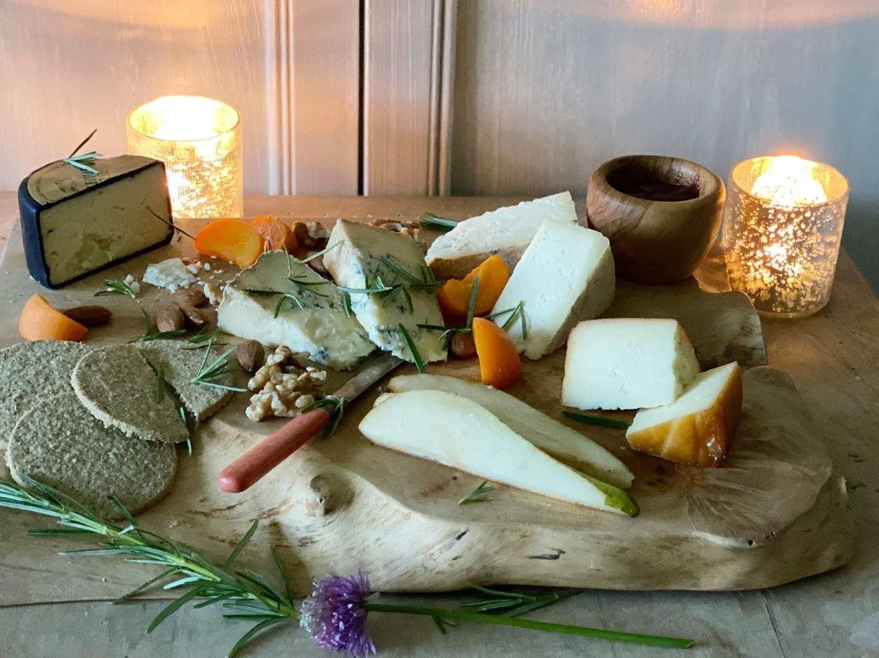 A cheese board with various cheeses, crackers, apricot slices, walnuts, and herbs, lit by two candles in glass holders and a small wooden bowl, on a wooden surface with a rustic background.