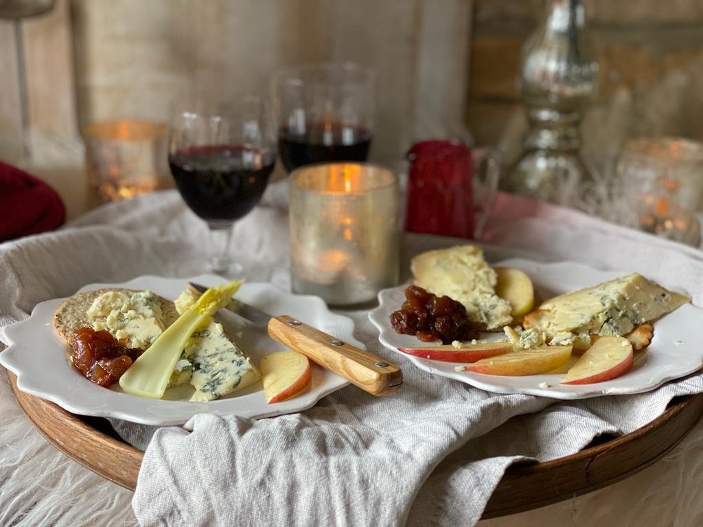 A cheese platter with apples, cheeses, and chutney on white plates, set on a wooden tray with three glasses of red wine in the background, surrounded by candles and rustic decor.