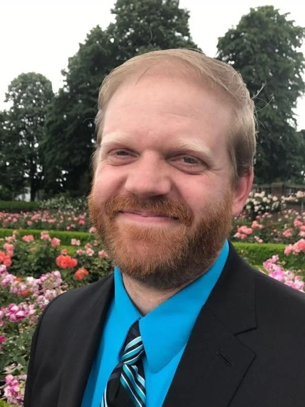 A man with a beard and light brown hair wearing a black suit, bright blue shirt, and striped tie, smiling outdoors in front of a garden with pink flowers and green trees.