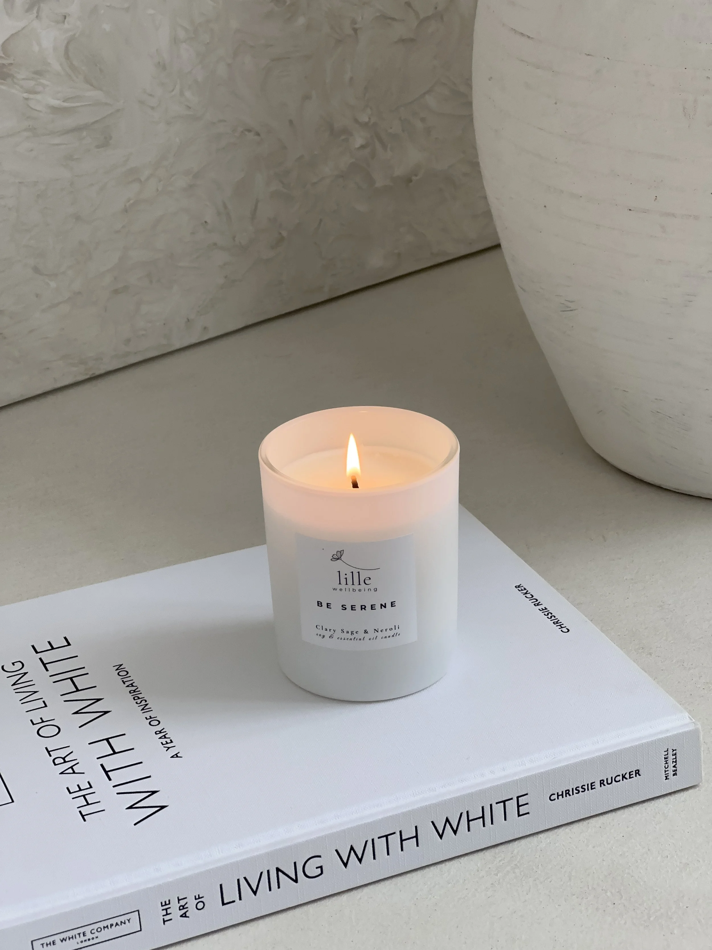 A white book titled 'LIVING WITH WHITE' with bold black text, placed on a white surface, with a lit white candle in front of it and off-white textured vases in the background.