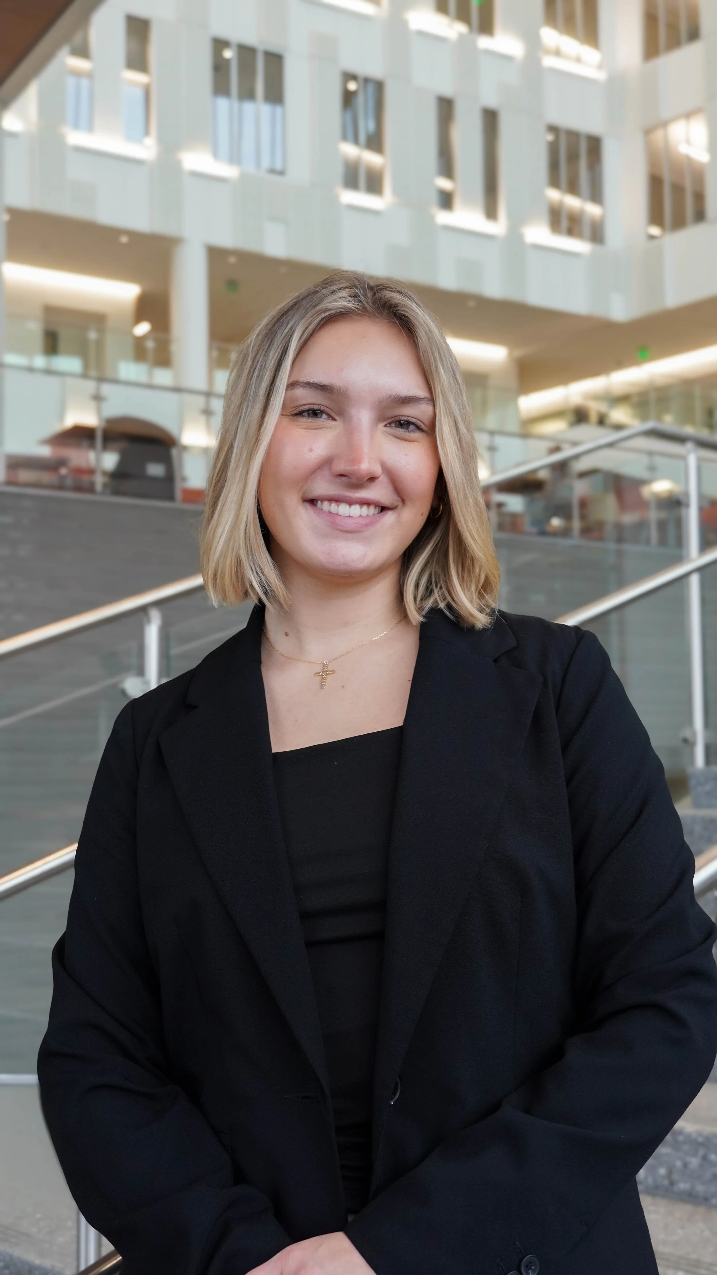 A young woman with blonde hair, wearing a black blazer and a cross necklace, smiling at the camera in a modern indoor setting with large windows and stairs.