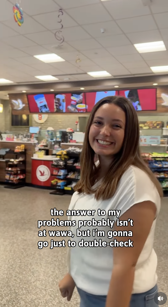 A young woman with long brown hair smiling inside a fast-food restaurant with digital menu boards and snack displays in the background.