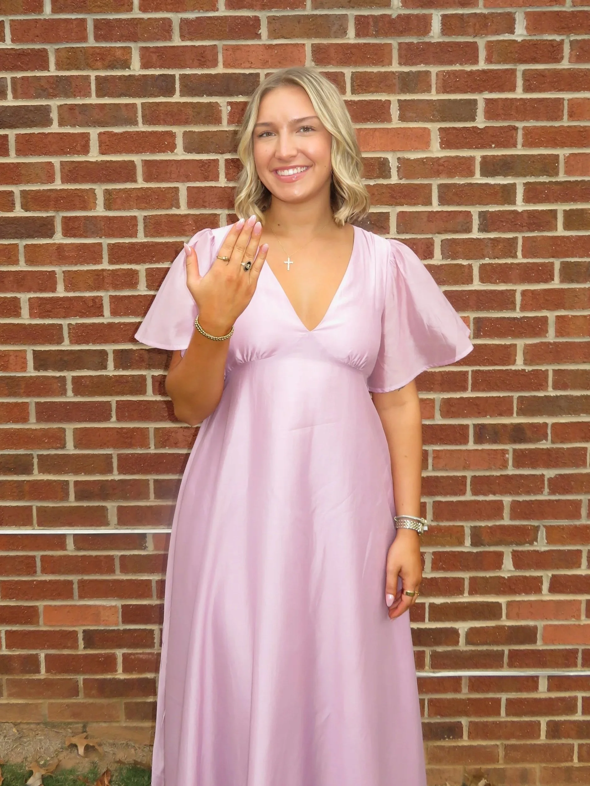 A smiling woman in a pink dress showing her left hand with a ring, standing against a brick wall.
