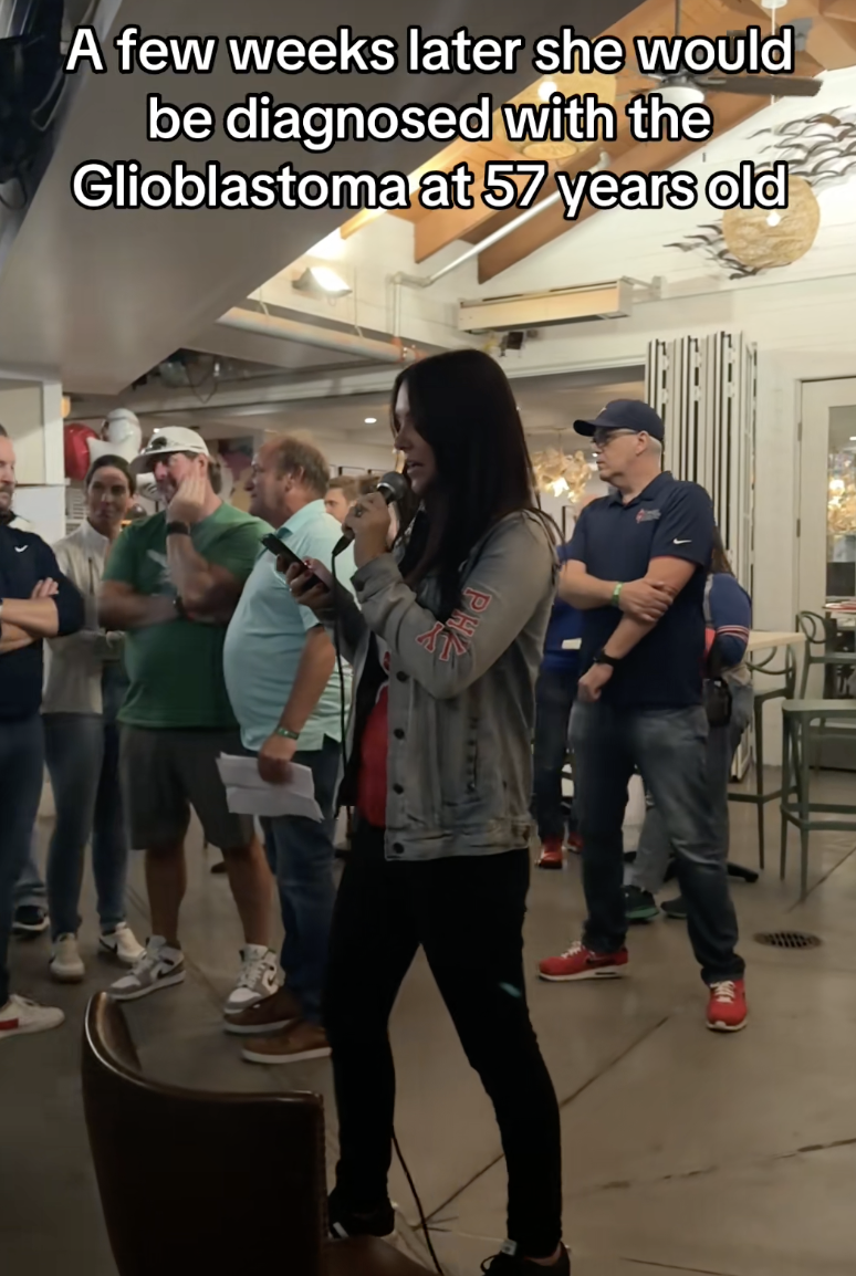 A woman speaks into a microphone while holding her phone in a room with a group of people gathered around her. The room has wooden beams on the ceiling and decorative lighting.