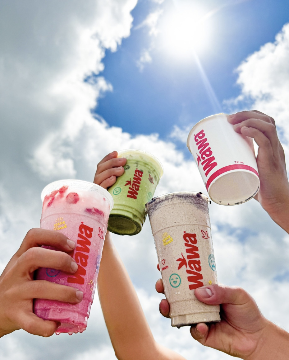 Four hands holding various flavored drinks in large cups up towards a blue sky with clouds and the sun.