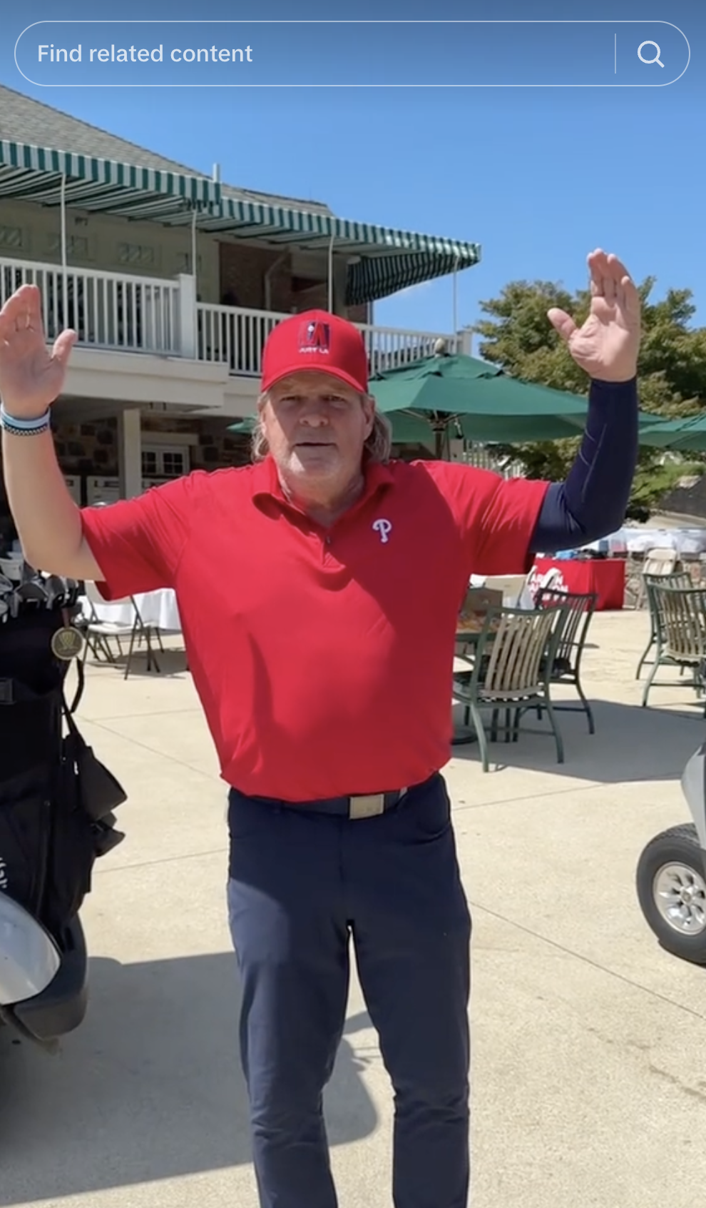 A man wearing a red baseball cap and red polo shirt with a white 'P' logo, raising his arms in an outdoor setting with tables, chairs, umbrellas, and a building with a balcony in the background.