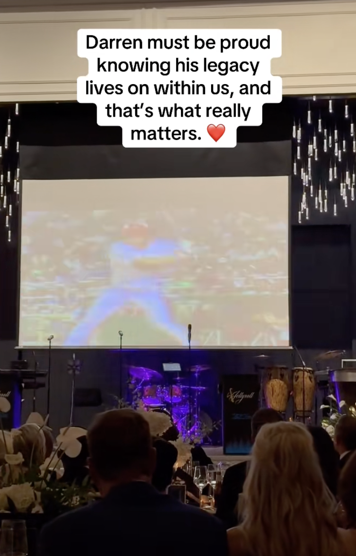 A large screen displaying a message about Darren's legacy at a formal event with a stage, musical instruments, and people seated in front. The message reads, "Darren must be proud knowing his legacy lives on within us, and that’s what really matters," with a red heart emoji.