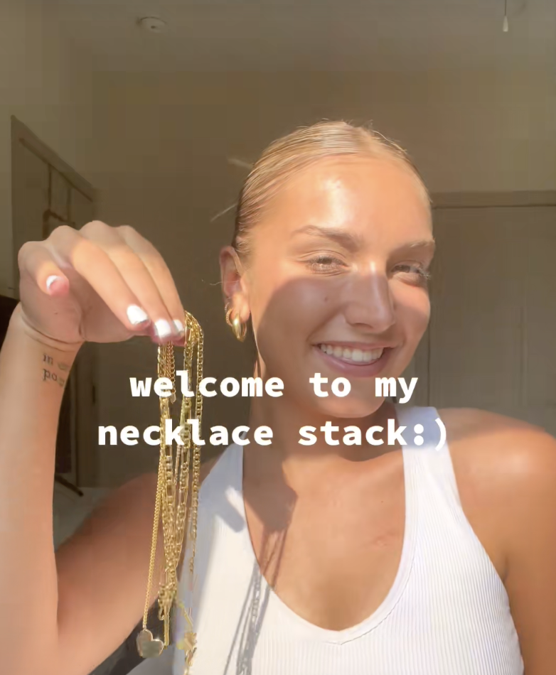 A woman with blonde hair tied back, smiling and holding up a gold necklace chain. She is wearing a white sleeveless top, and the background suggests she is indoors with warm lighting.