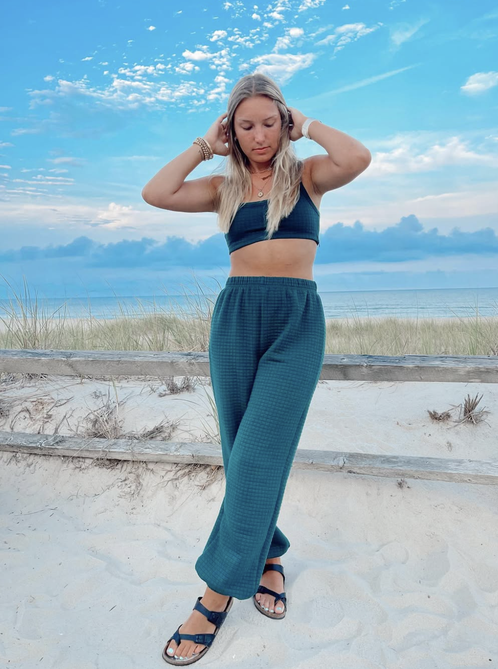 Young woman wearing a black sports bra, teal high-waisted pants, and sandals standing on a sandy beach with grass, wooden fence, and ocean in the background under a partly cloudy sky.