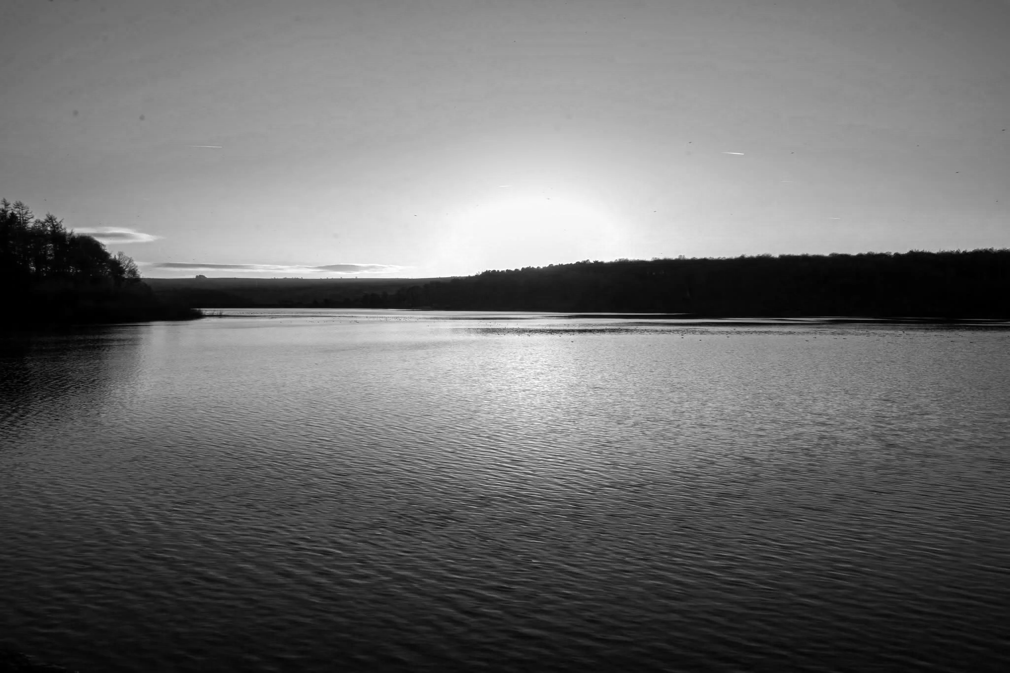 Black and white photo of a calm river with a slight ripple, with trees on the left and a hill in the distance across the water, under a bright sky.