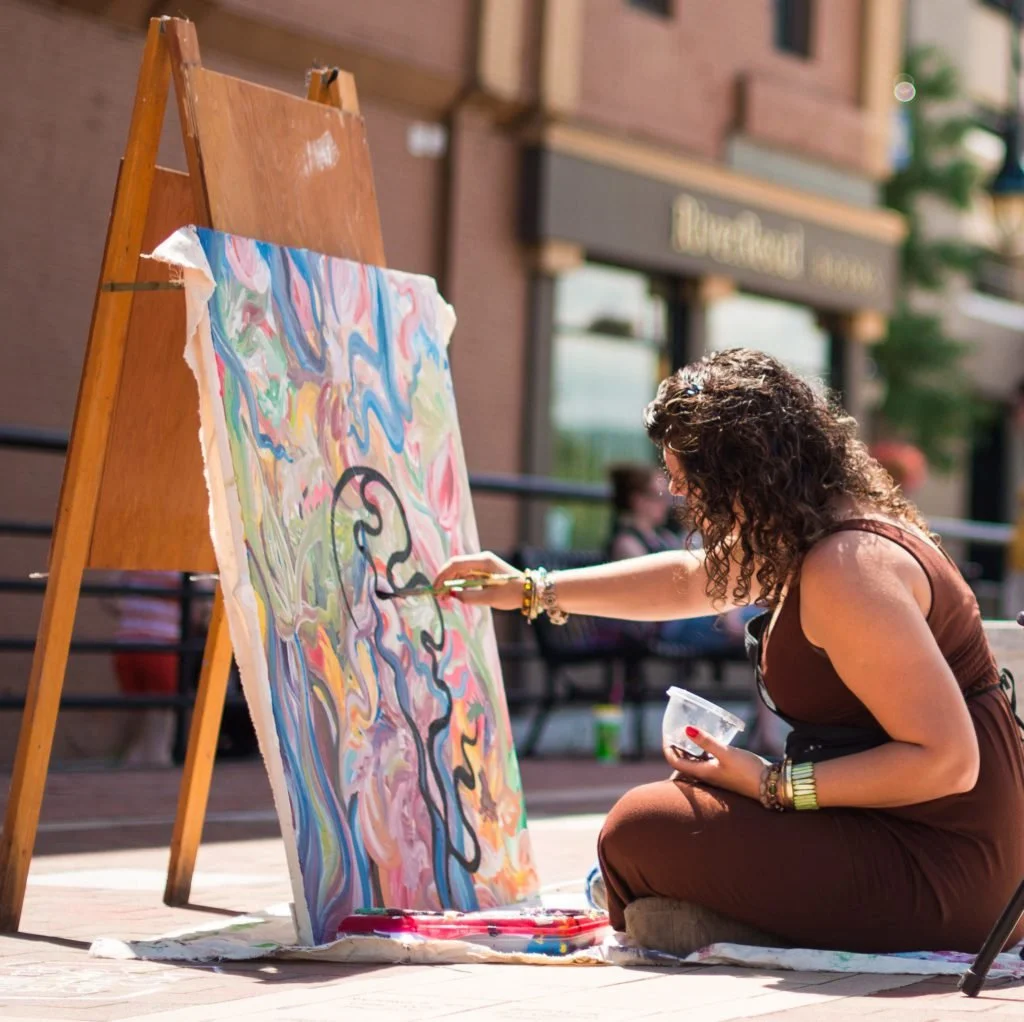 Artist sitting down, painting on an easel.