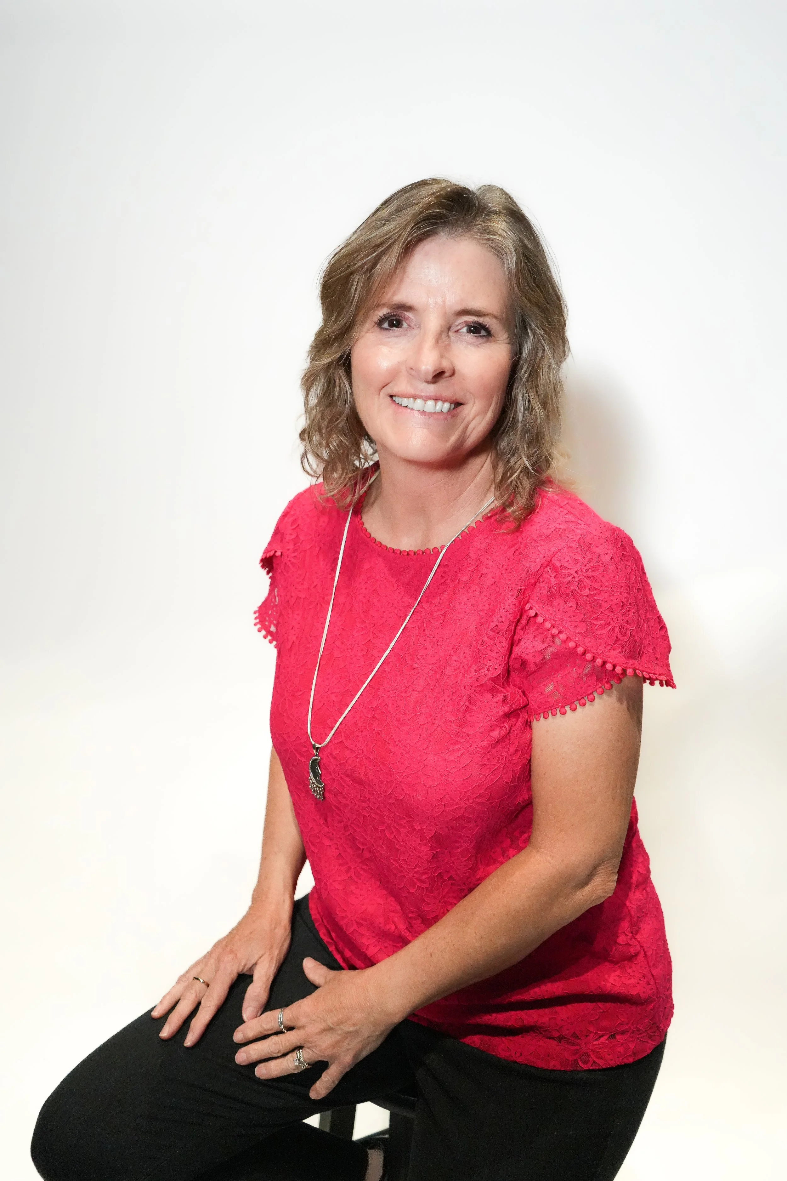 A woman with wavy, shoulder-length light brown hair and a warm smile, wearing a pink lace top and black pants, sitting on a stool against a plain white background.
