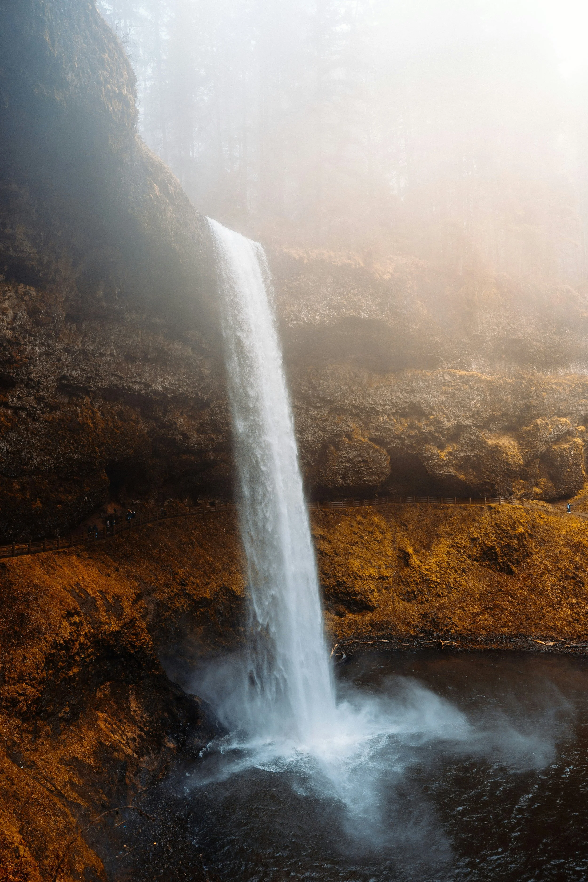 A tall waterfall cascading into a pool below, surrounded by mist and orange-brown rocky terrain.