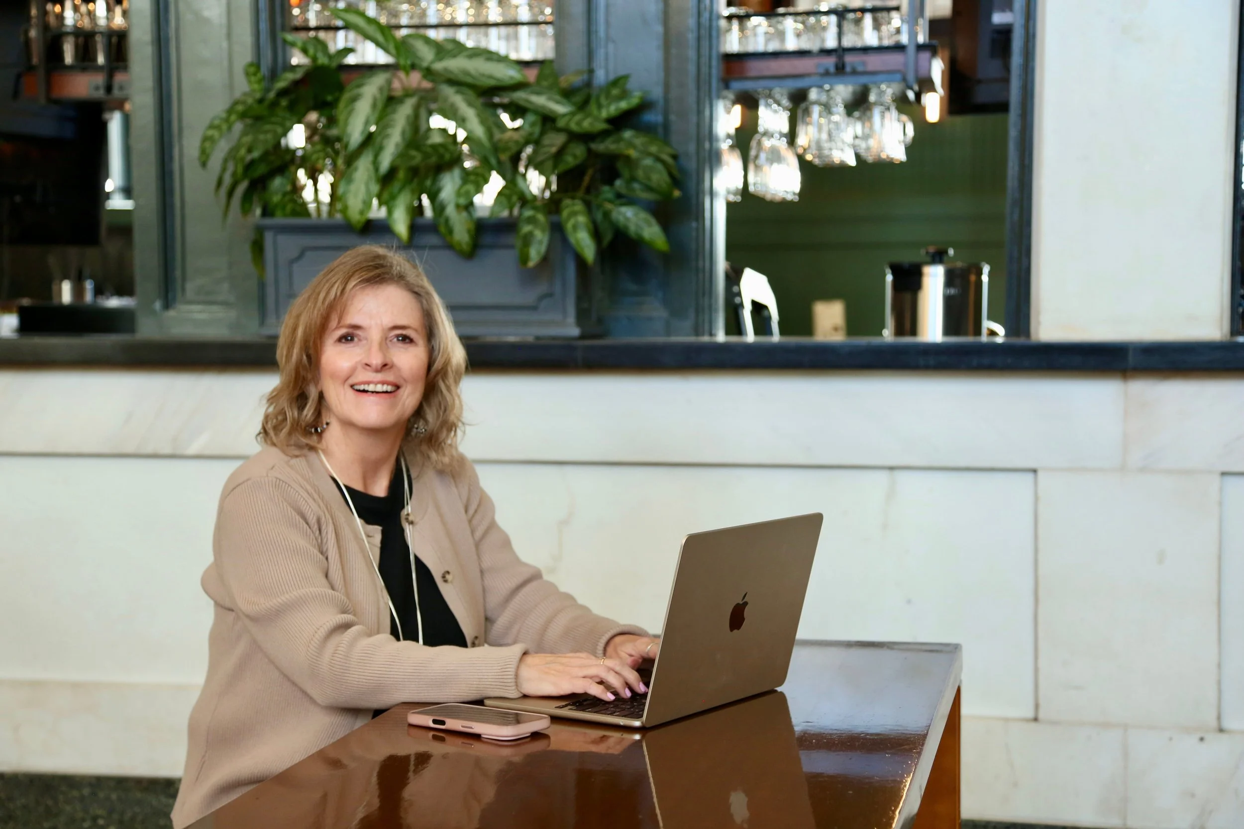 A woman with light brown, shoulder-length hair smiling while sitting at a table, working on a silver MacBook inside a cafe or restaurant.